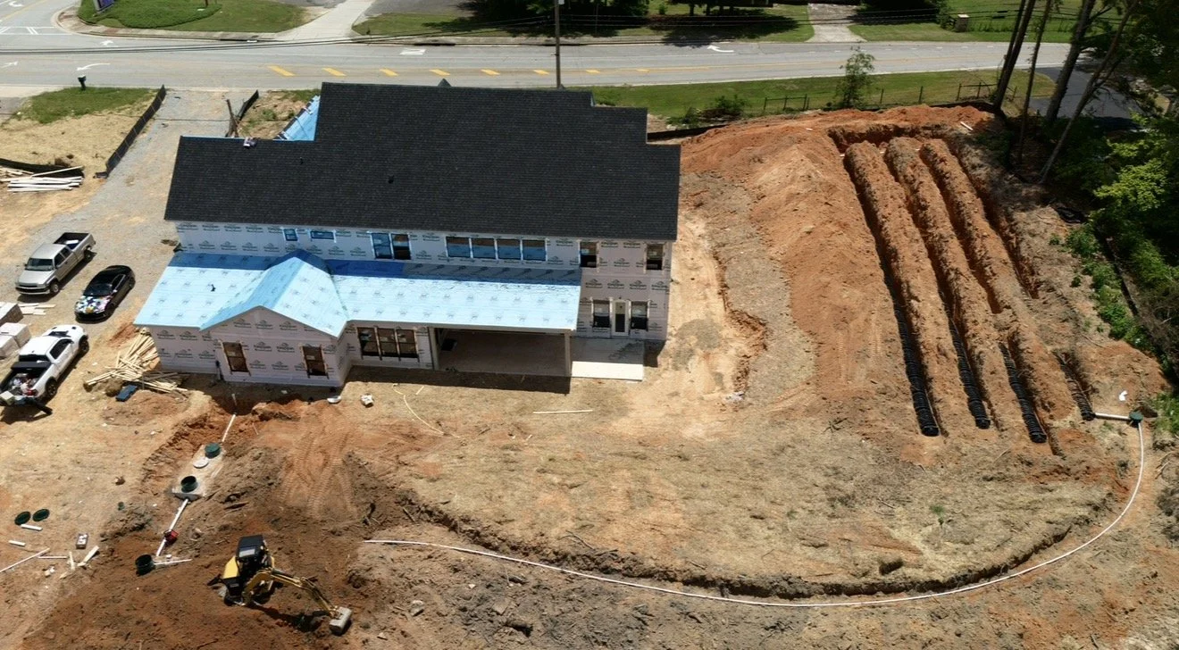A house under construction with surrounding construction site, multiple cars parked nearby, and a yard with several row trenches for underground piping or drainage.