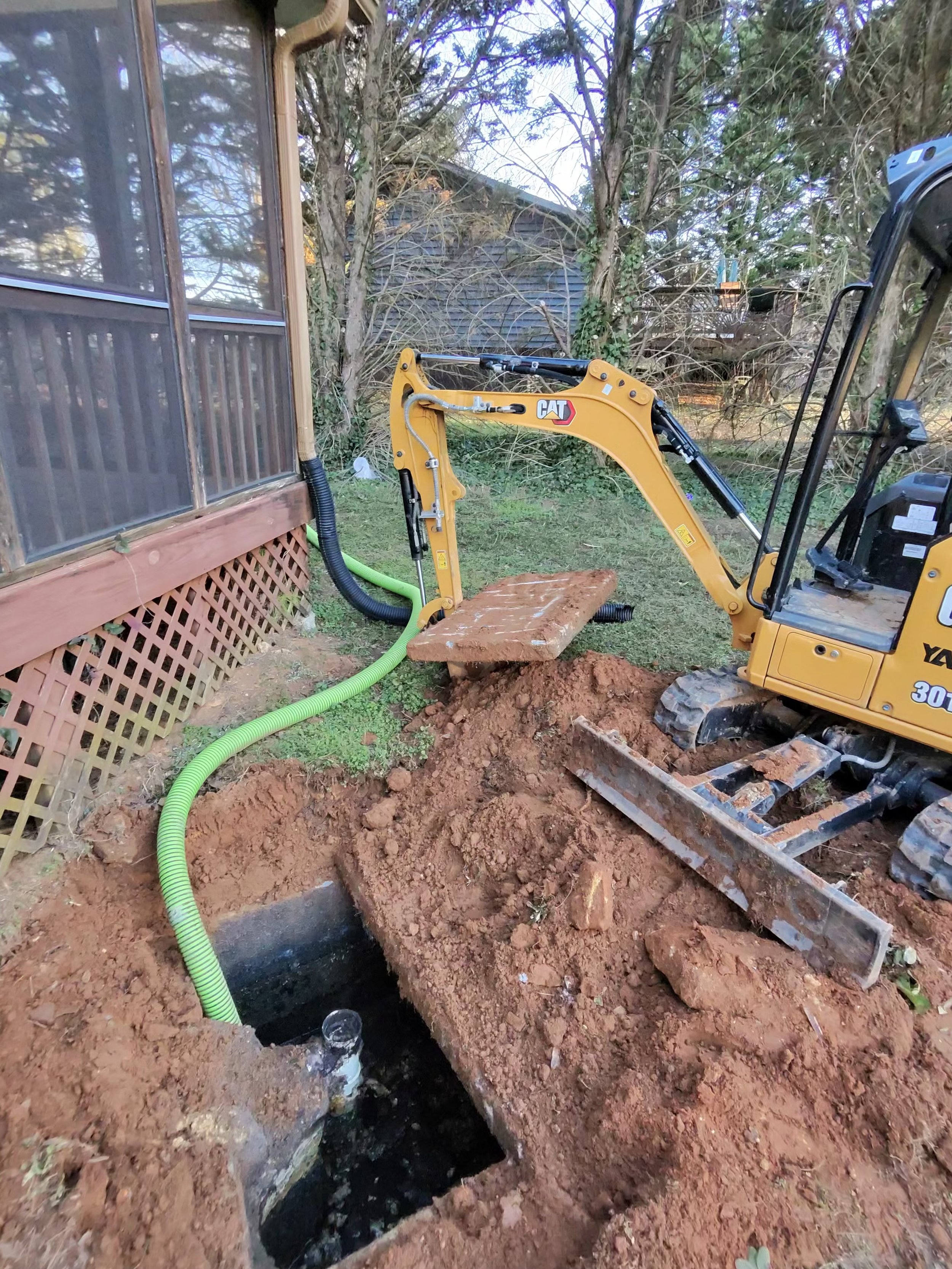 A small excavator digging a hole in the ground near a house with a screened porch. Green suction hoses for septic pumping are visible. Pumping out a septic tank with a PVC baffle.