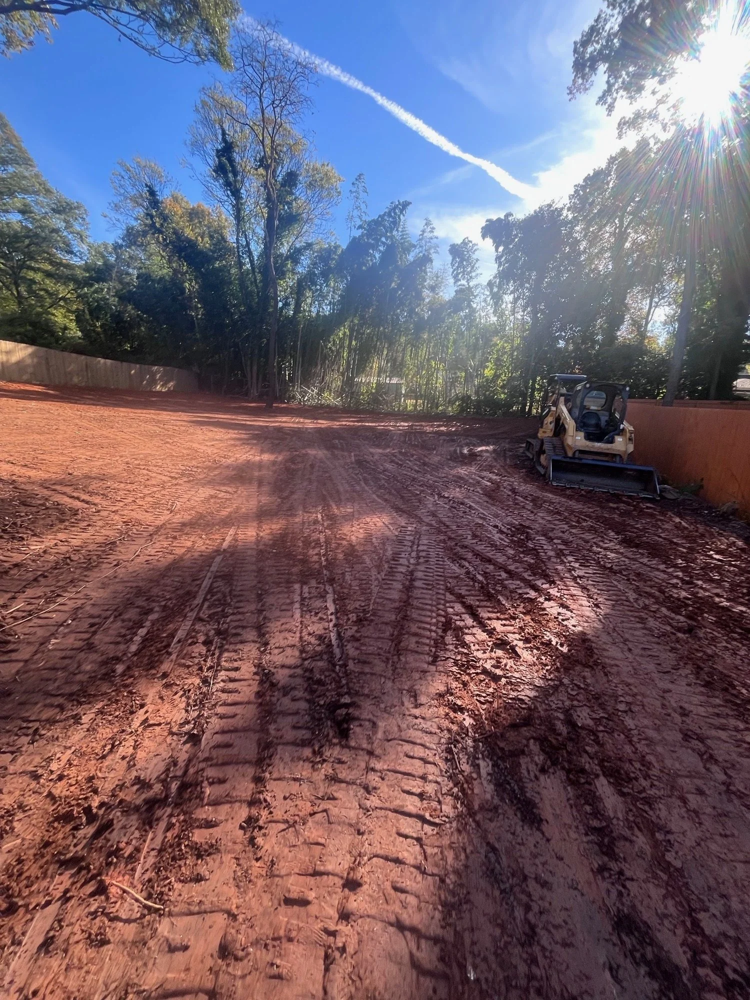 Construction site with red dirt ground, a small bulldozer to the right, tall trees and a clear blue sky with a contrail.