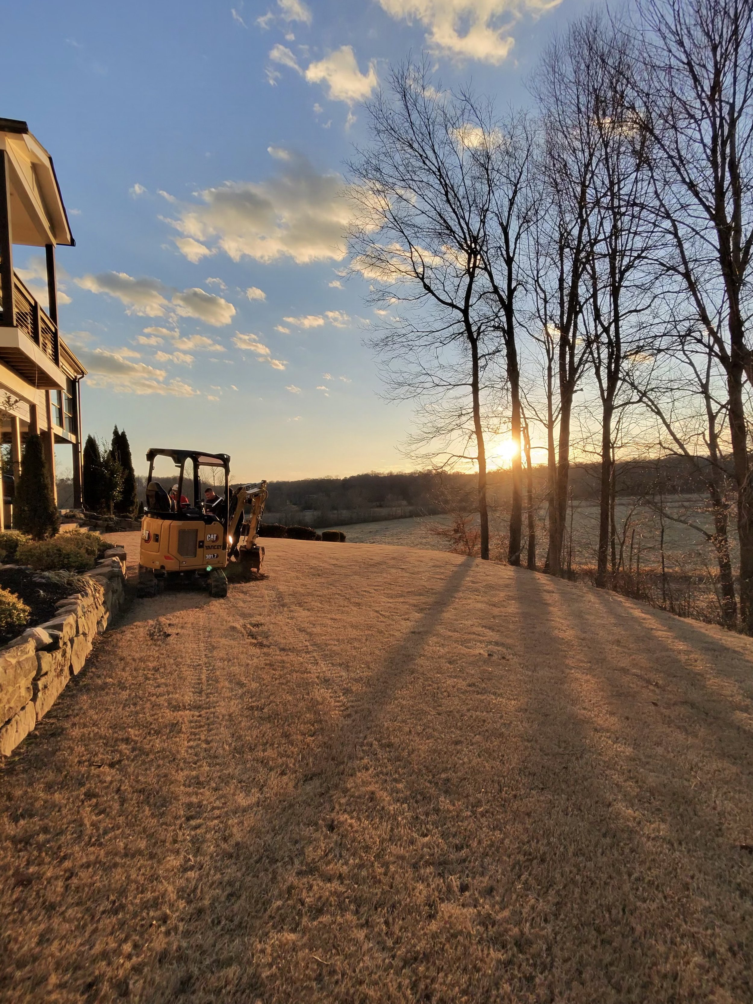 A sunset scene in a backyard with a small yellow excavator parked on a freshly cut grass lawn, leafless trees, a stone retaining wall, and a house on the left side. The sky has scattered clouds, and the sun is low on the horizon.