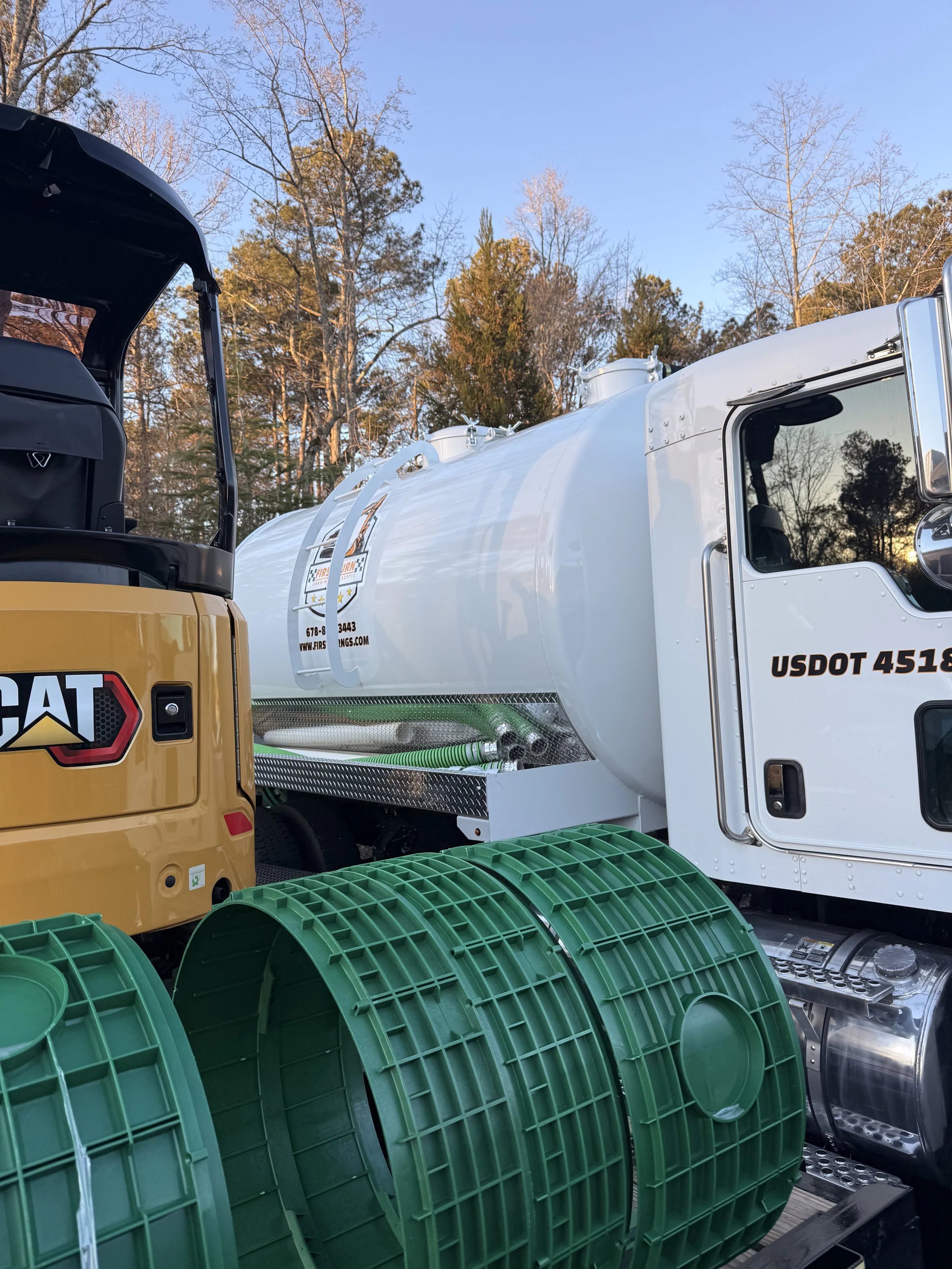 Close-up of a white tanker truck and a yellow construction vehicle with green hose reels, set outdoors with trees and a clear sky in the background.