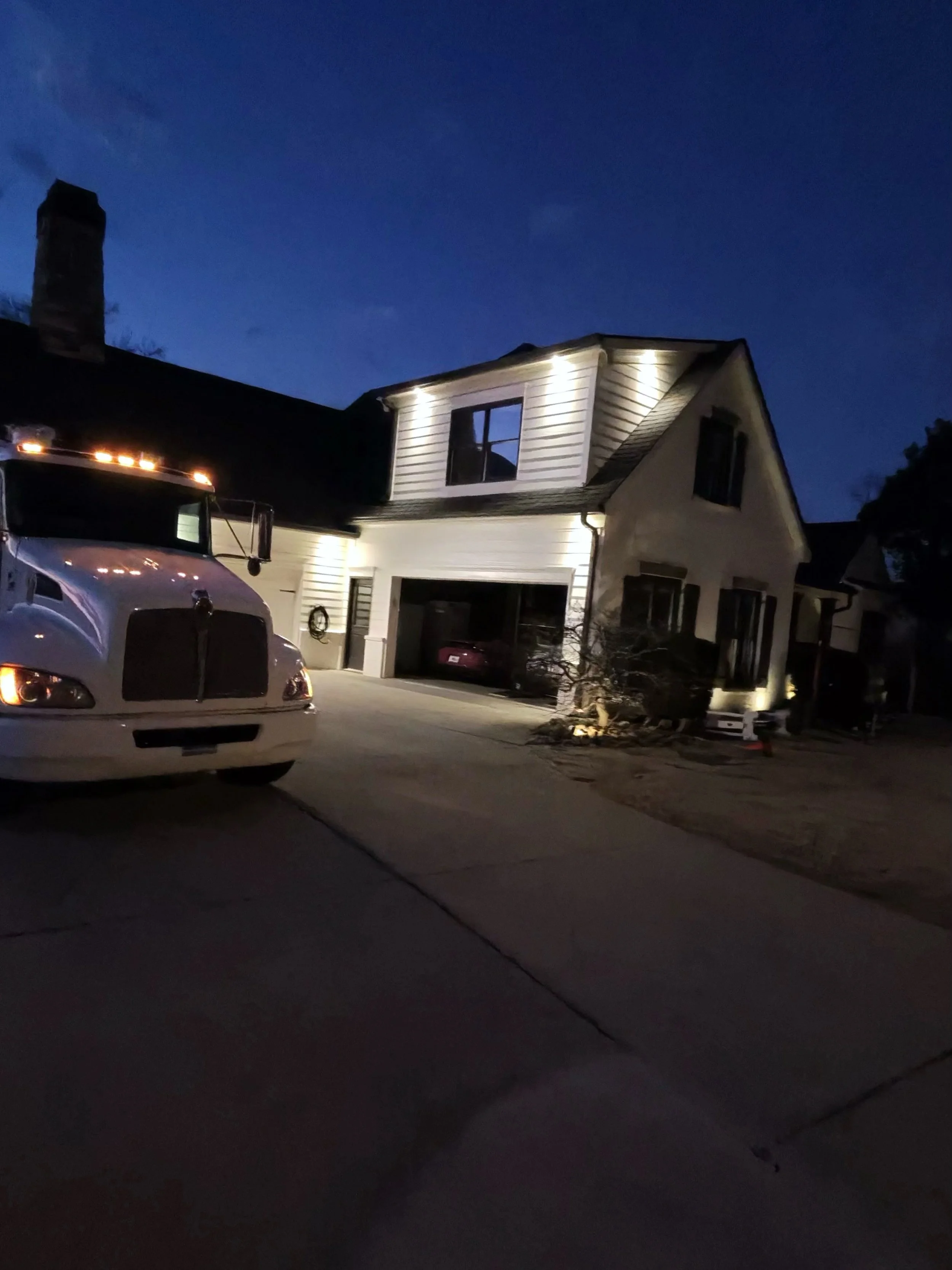 A two-story house with exterior lighting at night, a large white truck parked in driveway, and a garage with a car inside. The sky is dark with some clouds visible.