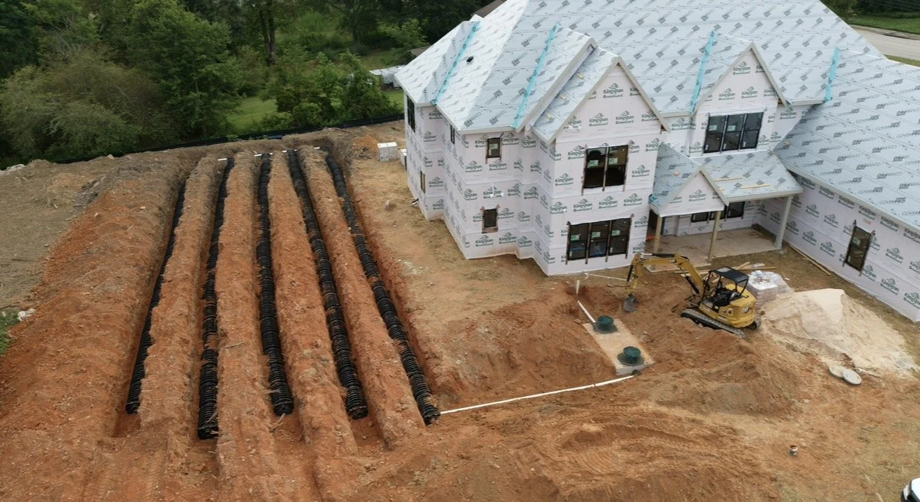 Construction site of a house with a white exterior and black windows, next to new septic drain fields with black plastic piping, surrounded by dirt and some greenery in the background.
