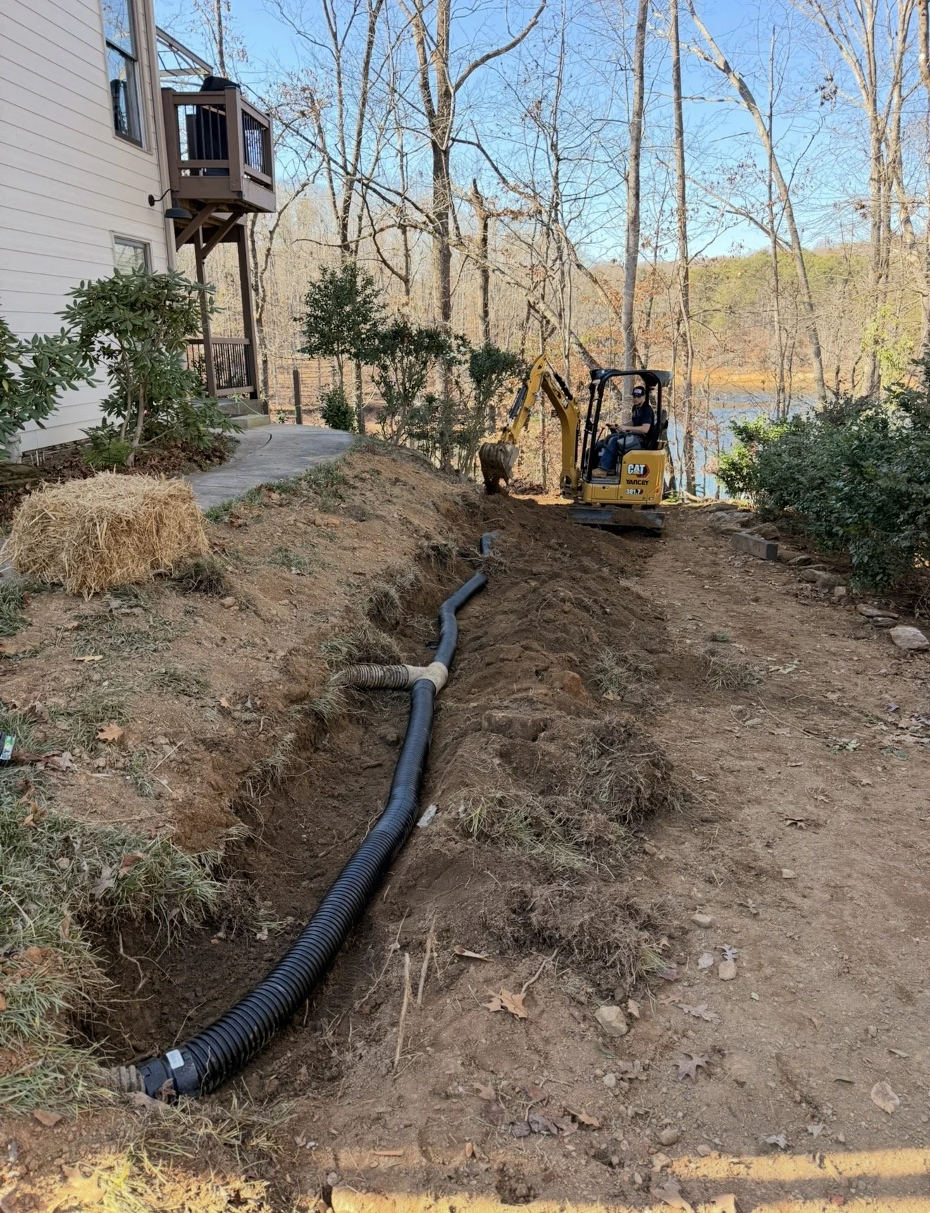 Construction worker operating a small excavator beside a lake, working on installing underground drainage pipes on a dirt pathway near a house.