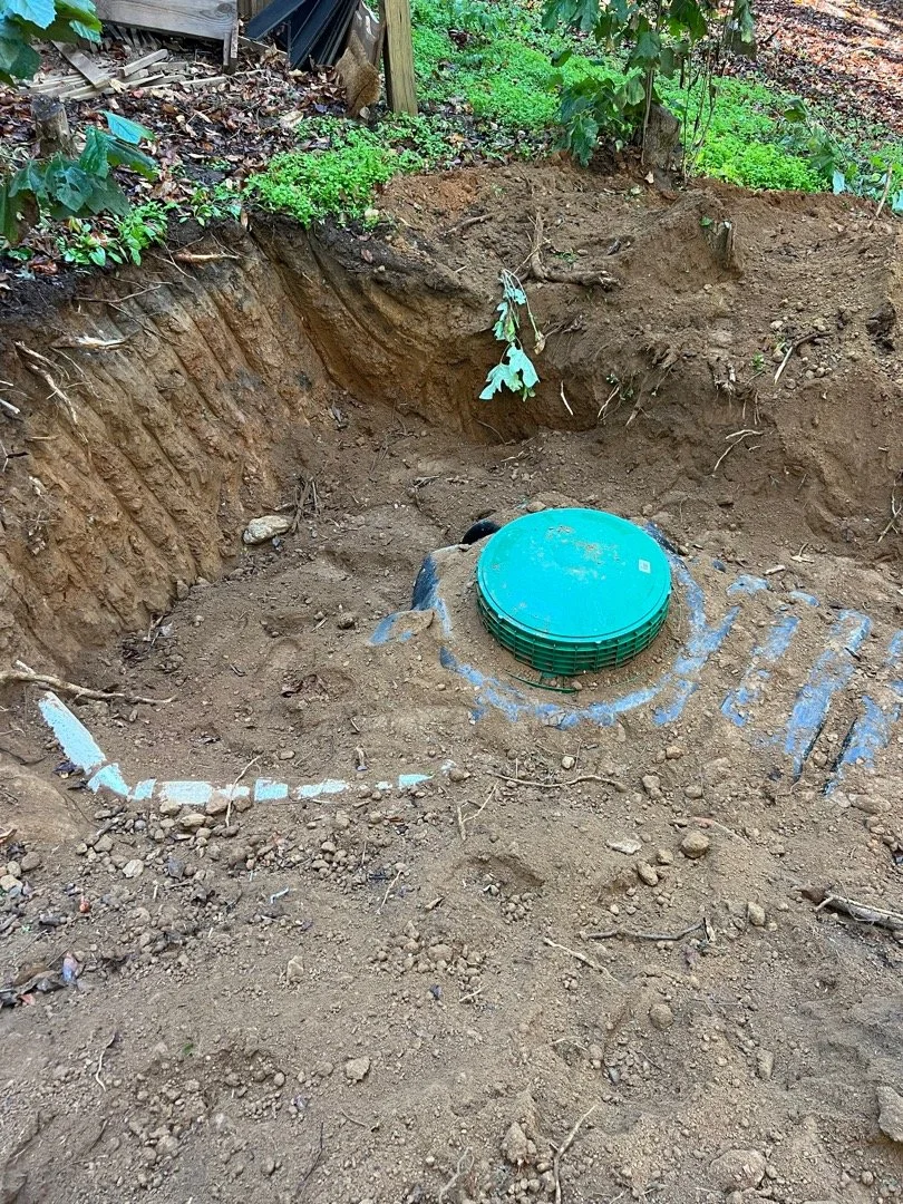 A dug hole in the ground with a green plastic sewer or utility cover installed at the bottom center. The surrounding area shows exposed soil and some plants and greenery around the edge of the excavation.