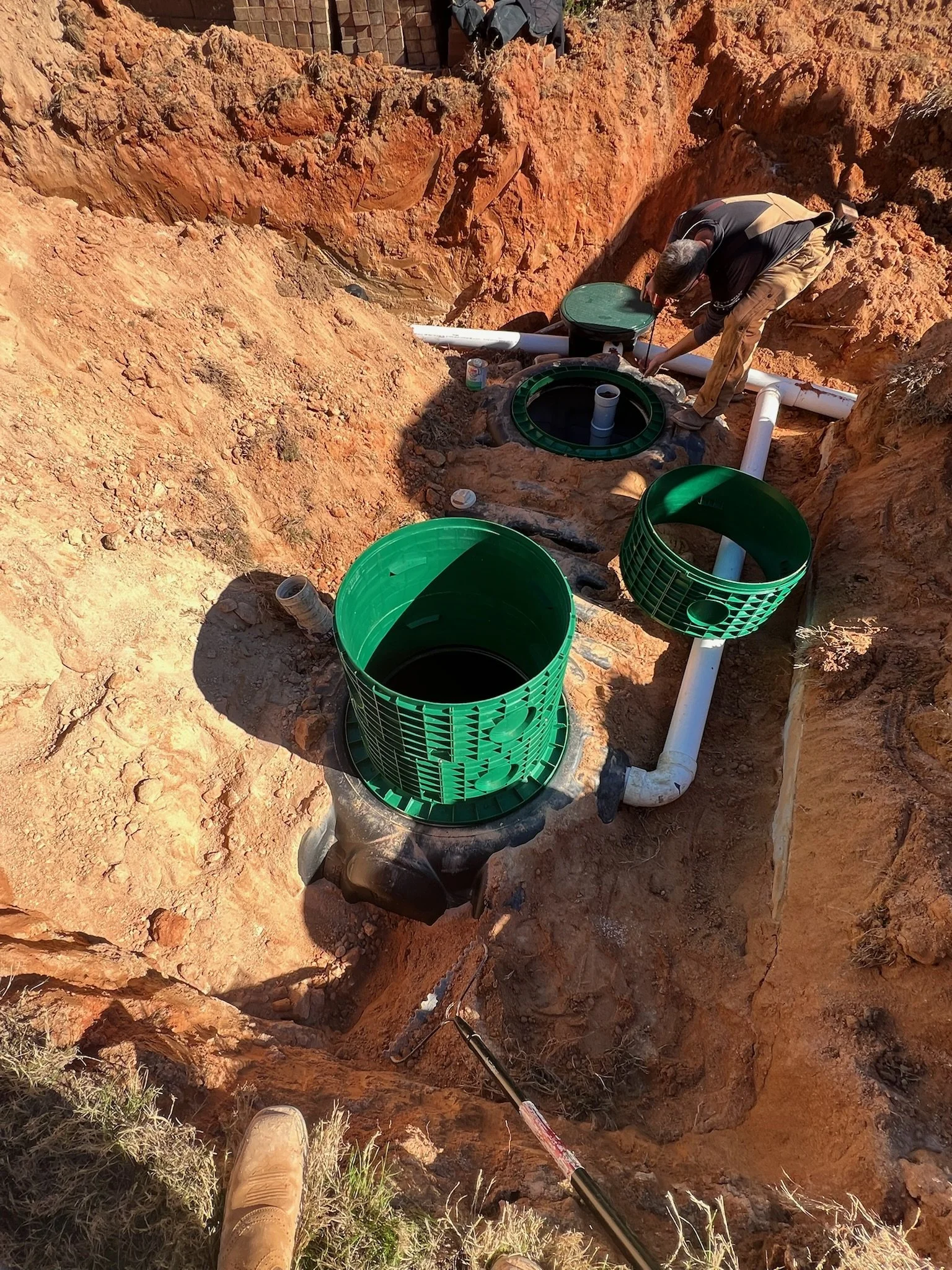 Worker installing underground plumbing with green septic tanks and white pipes in a construction trench.