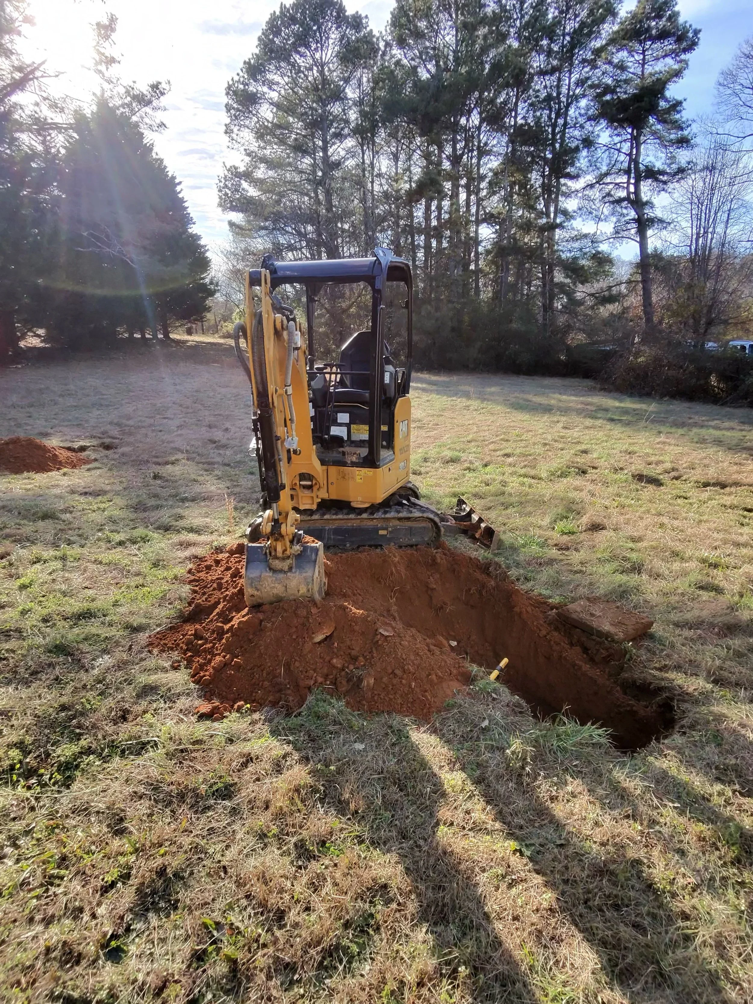 A small yellow excavator digging a trench in a grassy field during daytime.