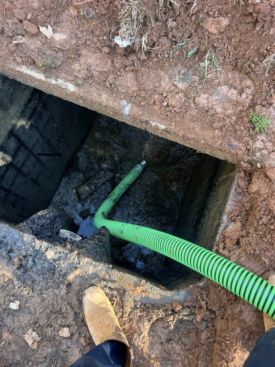 View of an open manhole with green corrugated pipe inside, surrounded by dirt, a shoe in the lower left corner, and some small plants.