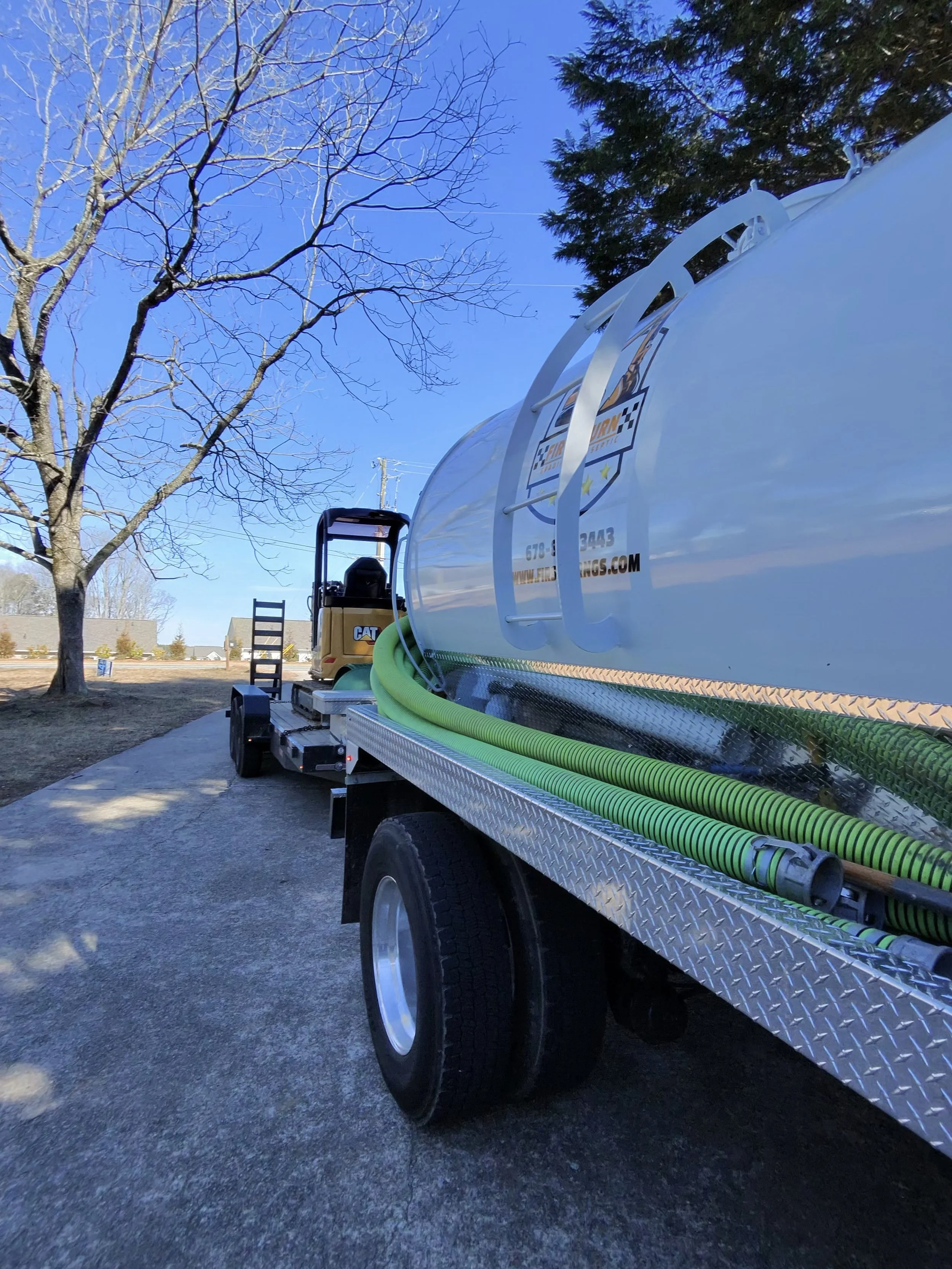 Close-up of a large white tanker truck with green hoses attached, parked on a paved surface near a tree with bare branches and a clear blue sky in the background.