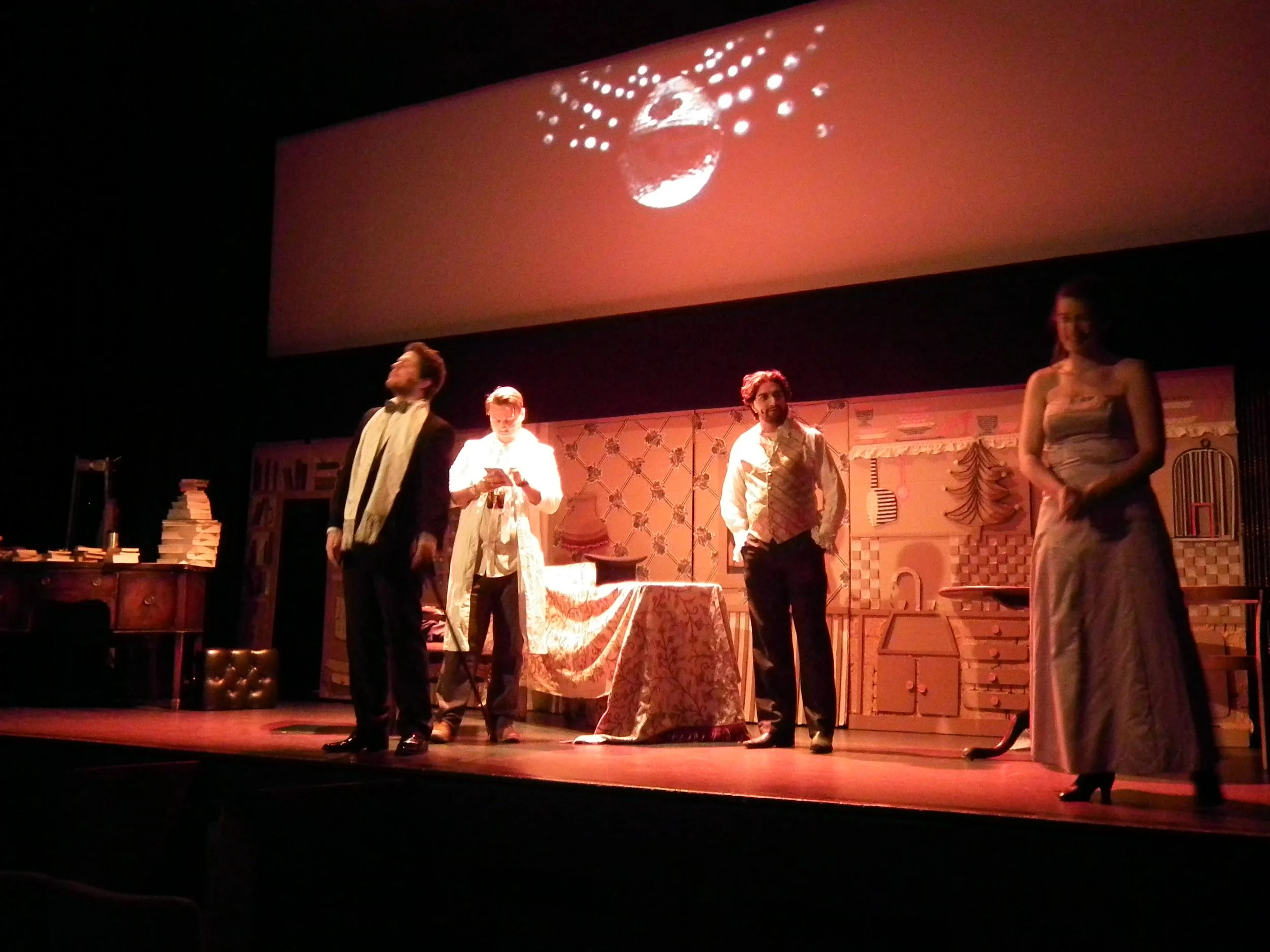 Theater stage with four actors performing a scene. The background has shelves filled with books and various props. A large screen above displays a disco ball.