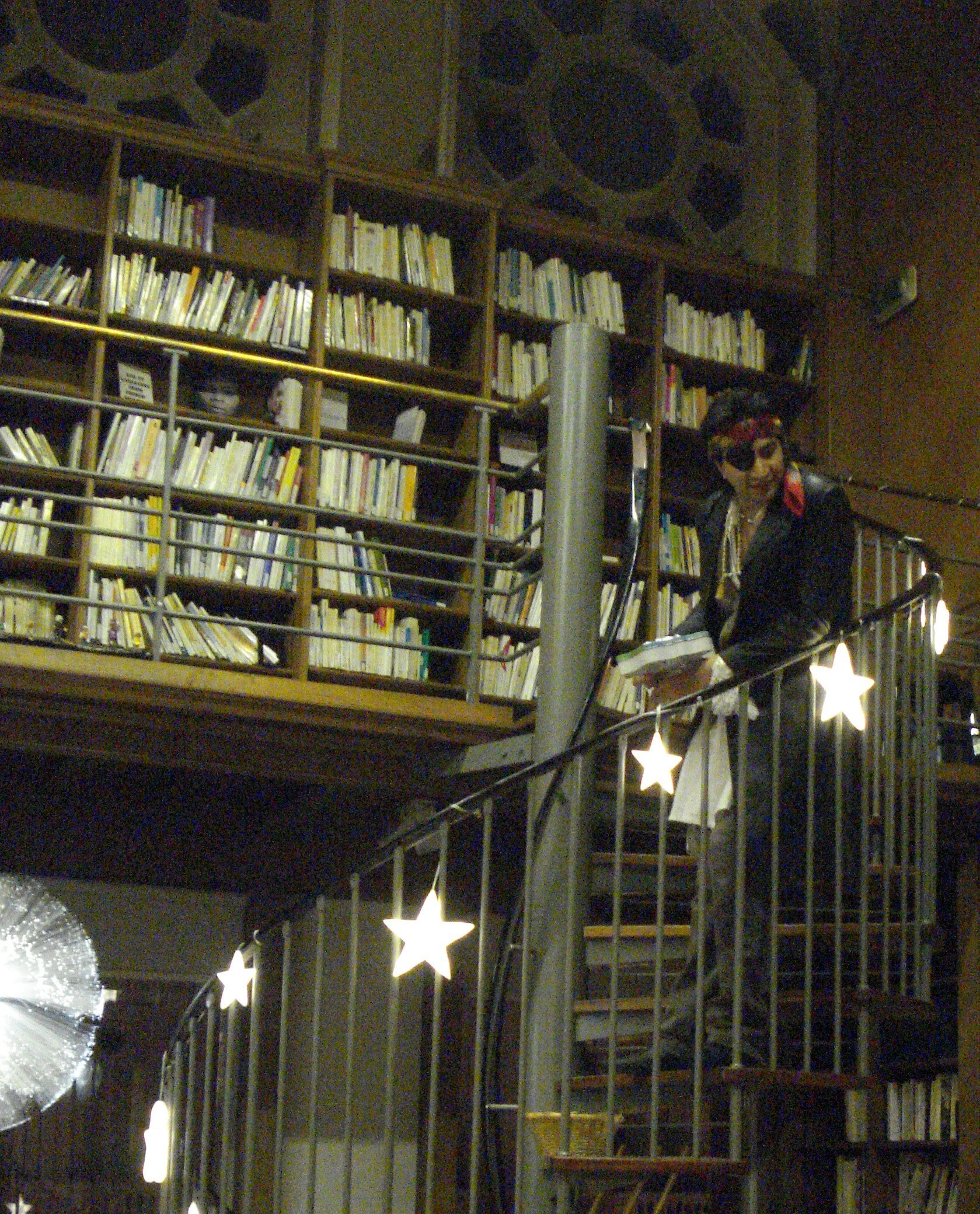 Une personne vêtue d'une veste noire et d'un chapeau coloré se tient sur un escalier en colimaçon à l'intérieur d'une bibliothèque. À l'arrière-plan, on aperçoit de hautes étagères en bois remplies de livres et des guirlandes lumineuses.
