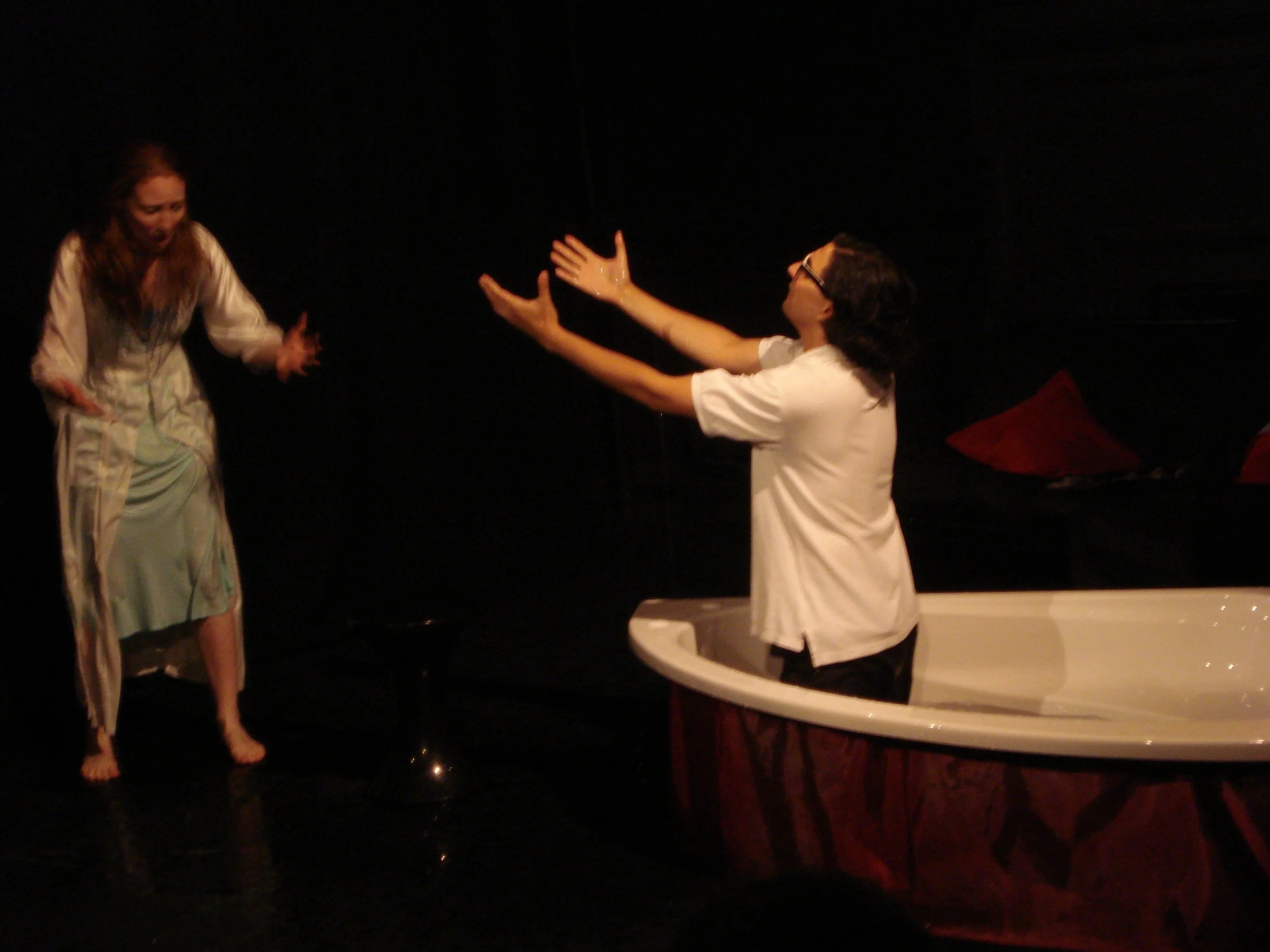 Two people onstage; a woman is standing, visibly distressed, and a man is in a bathtub, reaching out with his arms.
