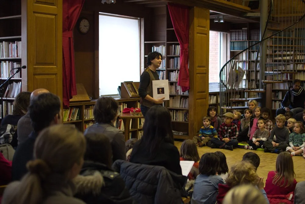 A man standing in front of a group of children and adults, holding a book and giving a performance. The children are seated on the floor, attentively listening, with bookshelves around them.
