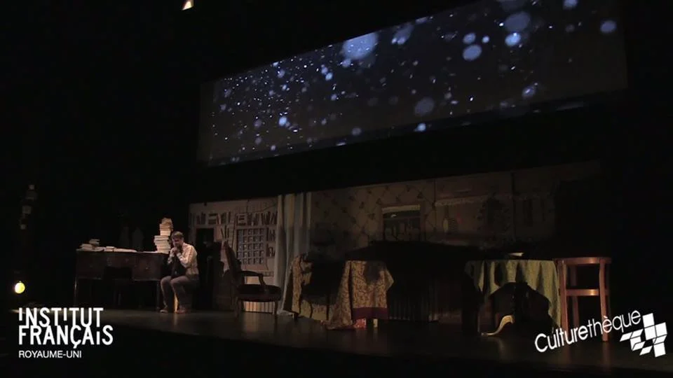 Stage scene with a person sitting on a chair and a table with a pile of books and papers, background decorated with curtains and furniture, and a large screen above showing an animation of falling snow or stars