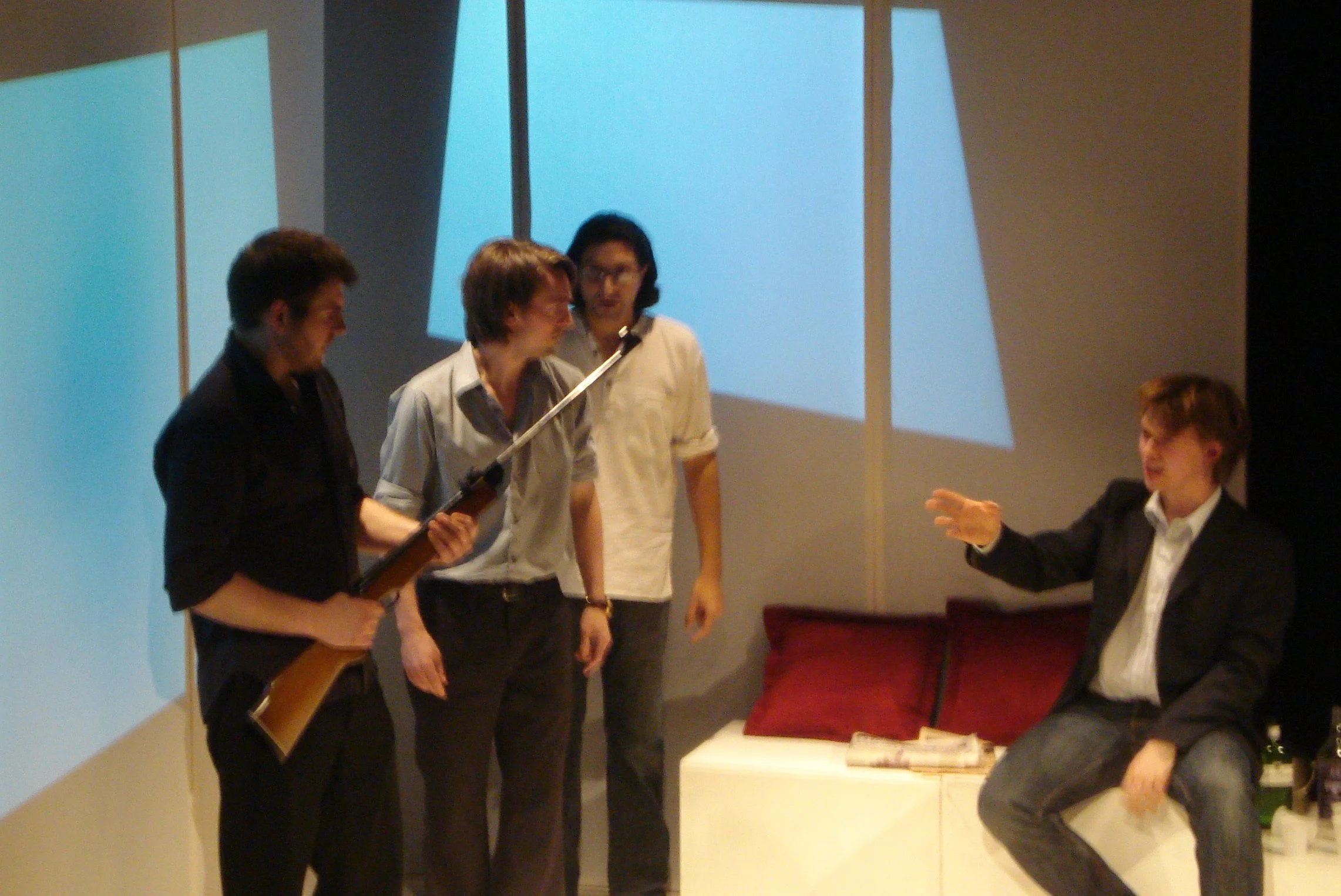 Four people in a room, three standing and one sitting, engaged in conversation. The person sitting is gesturing with their hand, and the three standing are looking at them. The person on the left is holding a rifle.