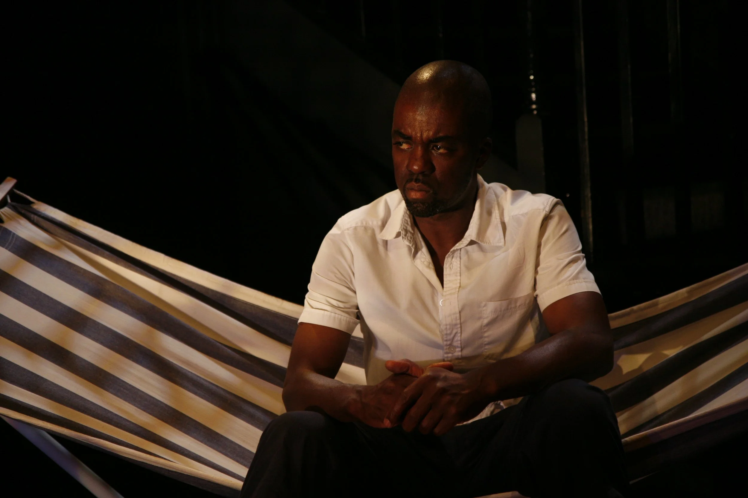 A man sitting on a striped hammock onstage, wearing a white shirt and black pants, with a serious expression.