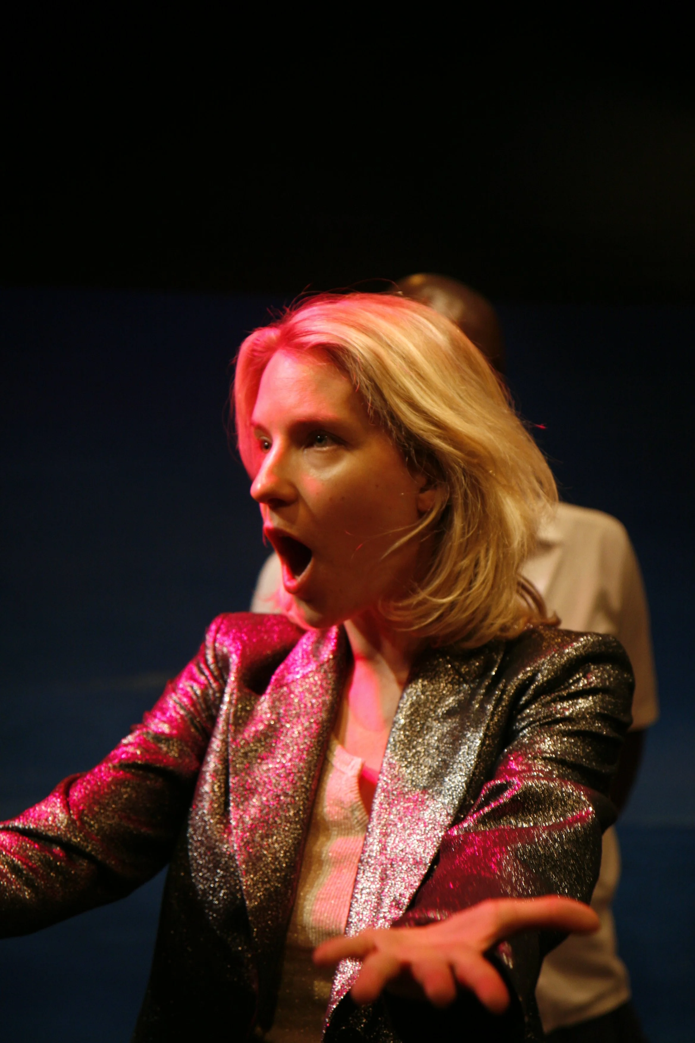 A woman wearing a shiny, metallic blazer, expressing surprise, with her mouth open and eyes wide, on stage with dark background.