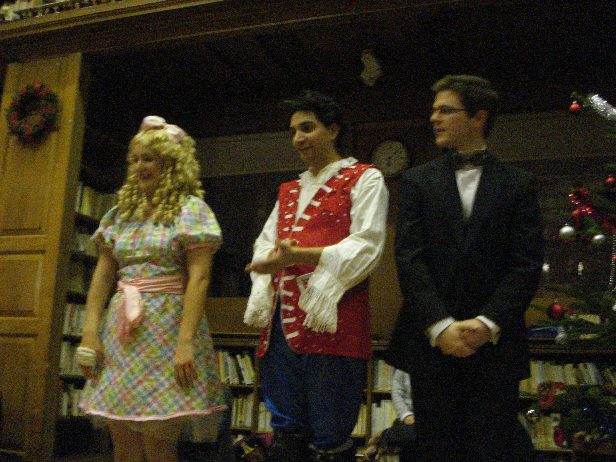 Trois personnes déguisées se tiennent devant un sapin de Noël et des étagères, participant à un spectacle de Noël.