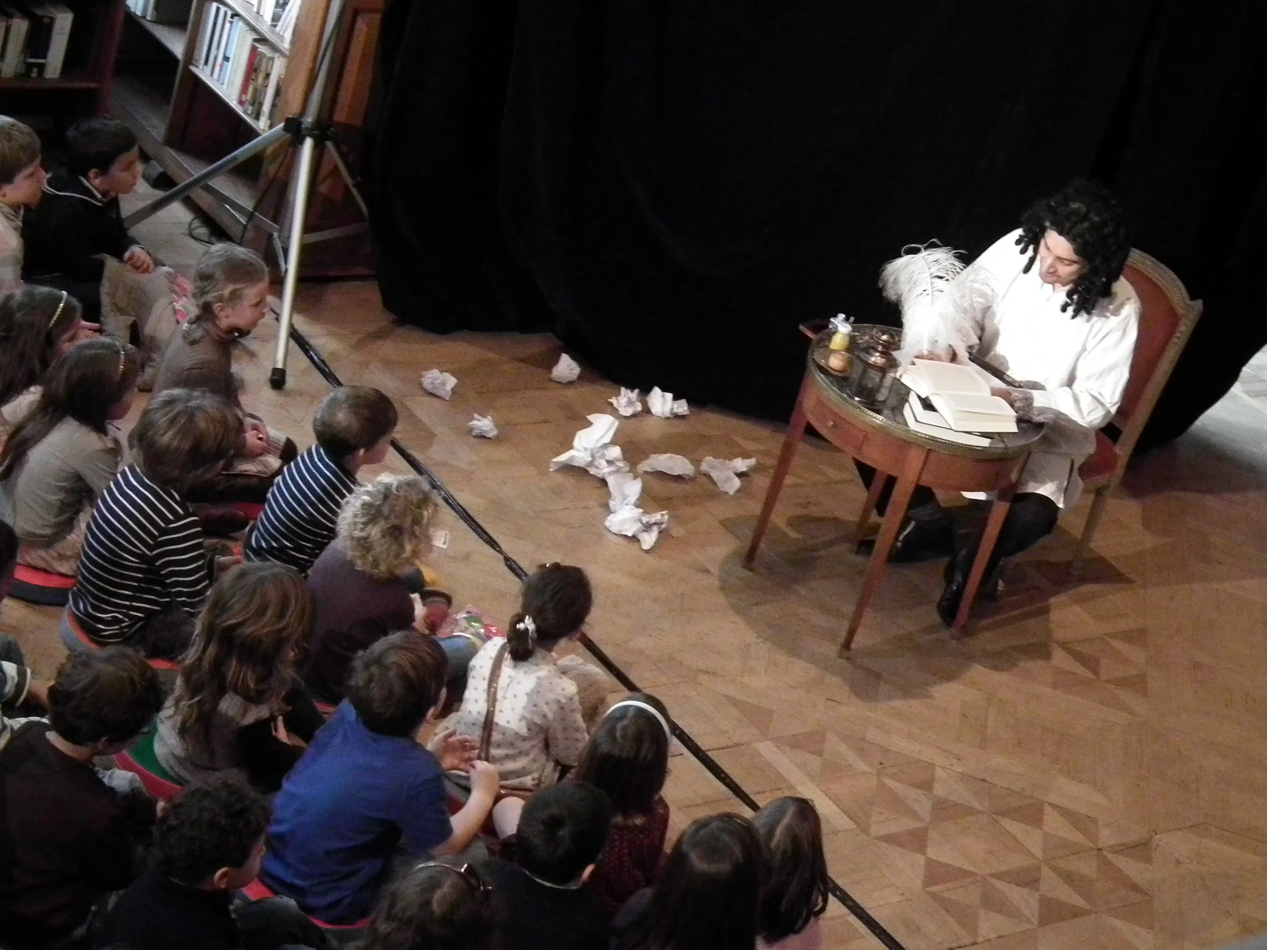 A person dressed in a wig as Charles Perrault reading to a group of children seated on the floor in a library.