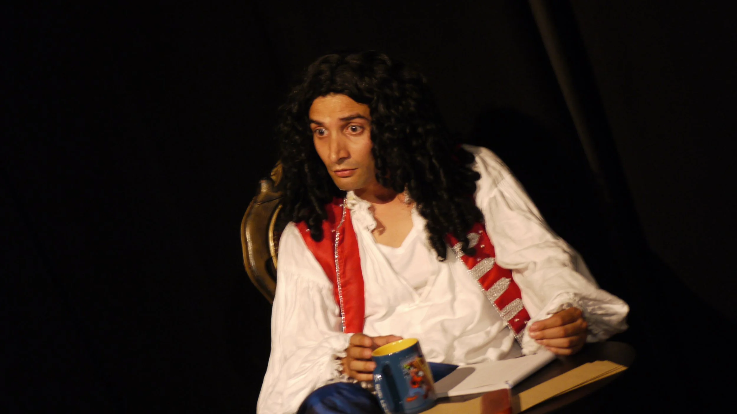 A man with a long curly black wig, dressed in a white ruffled shirt and red and silver vest, sitting onstage, holding a blue mug with a cartoon character, with a surprised expression.