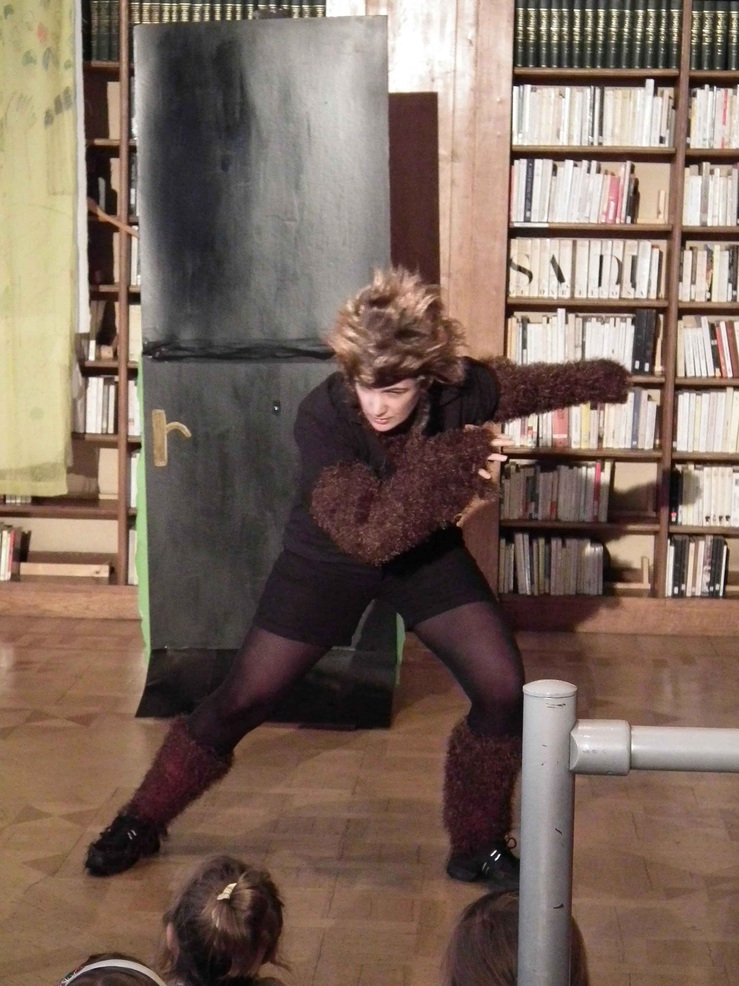 A woman in a wolf costume performs a theatrical act in front of an audience in a library setting, with children watching in the foreground.