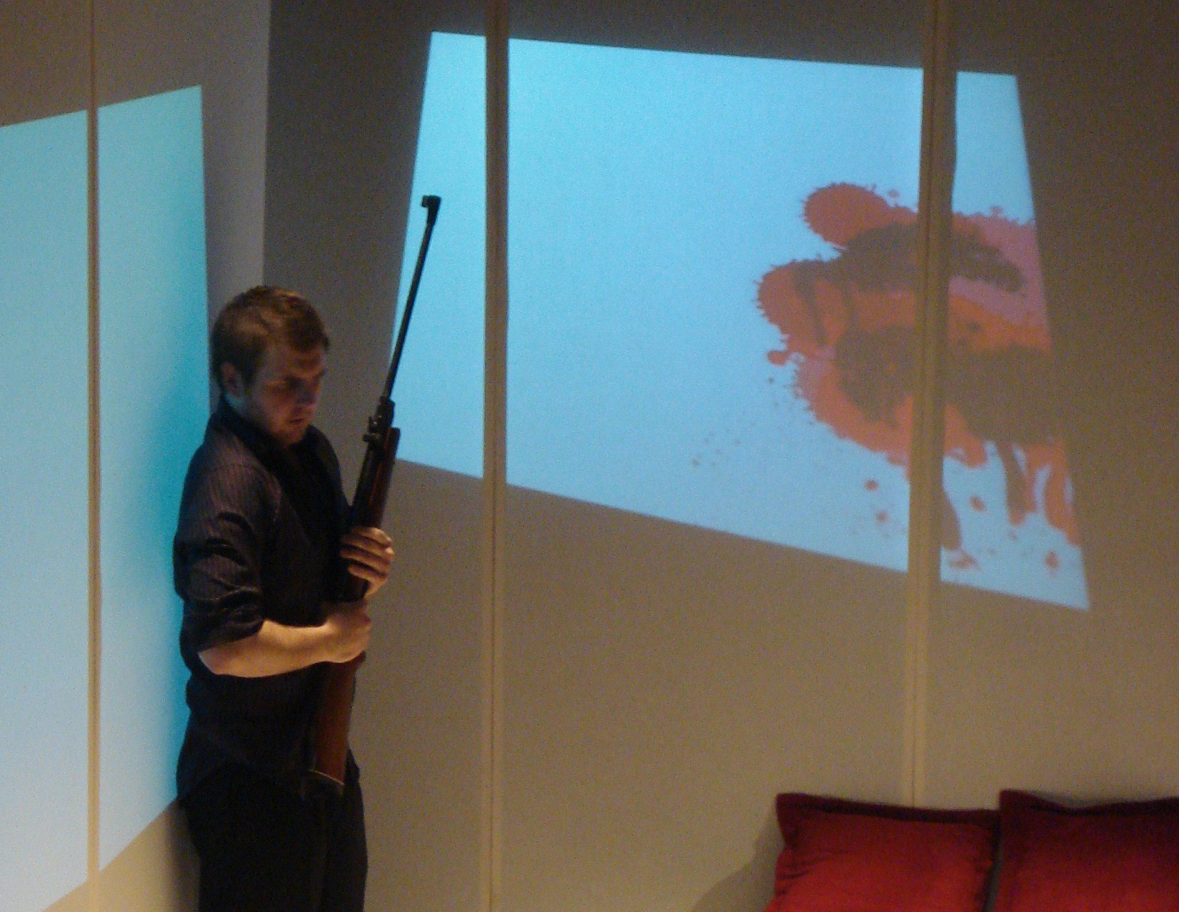 A person standing onstage, holding a rifle, with a large projected image of a red and black inkblot on a screen or wall behind them.