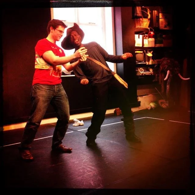 Two young men dressed as characters from’ the cat in boots’ are performing in a rehearsal room with props and equipment