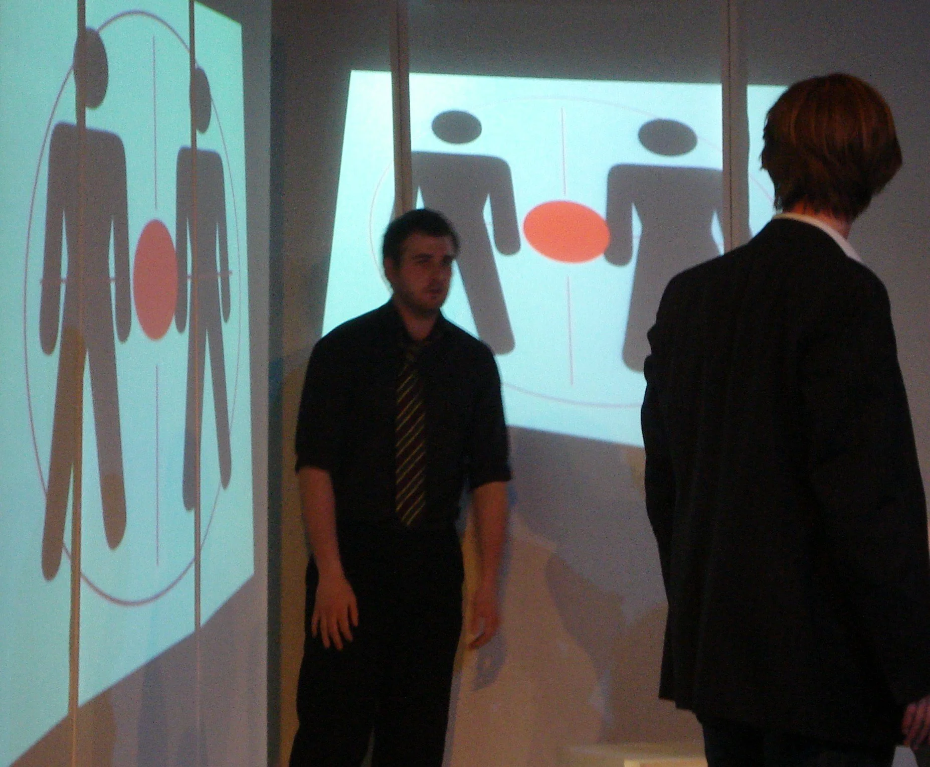 Two men stand in front of a projection screen displaying a diagram of two human figures facing each other, with an red circle between them