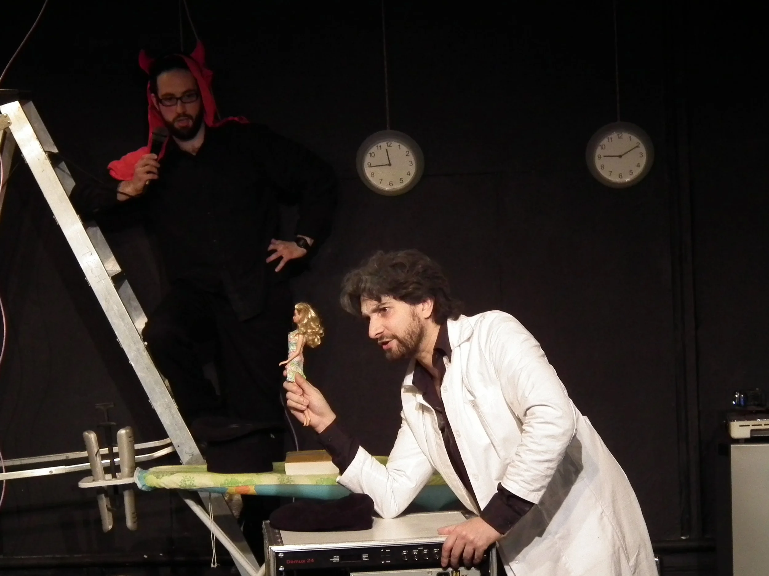 Two men performing in a stage play: one man dressed as a scientist holding a doll, another man with glasses dressed as a devil standing on a ladder with a microphone.