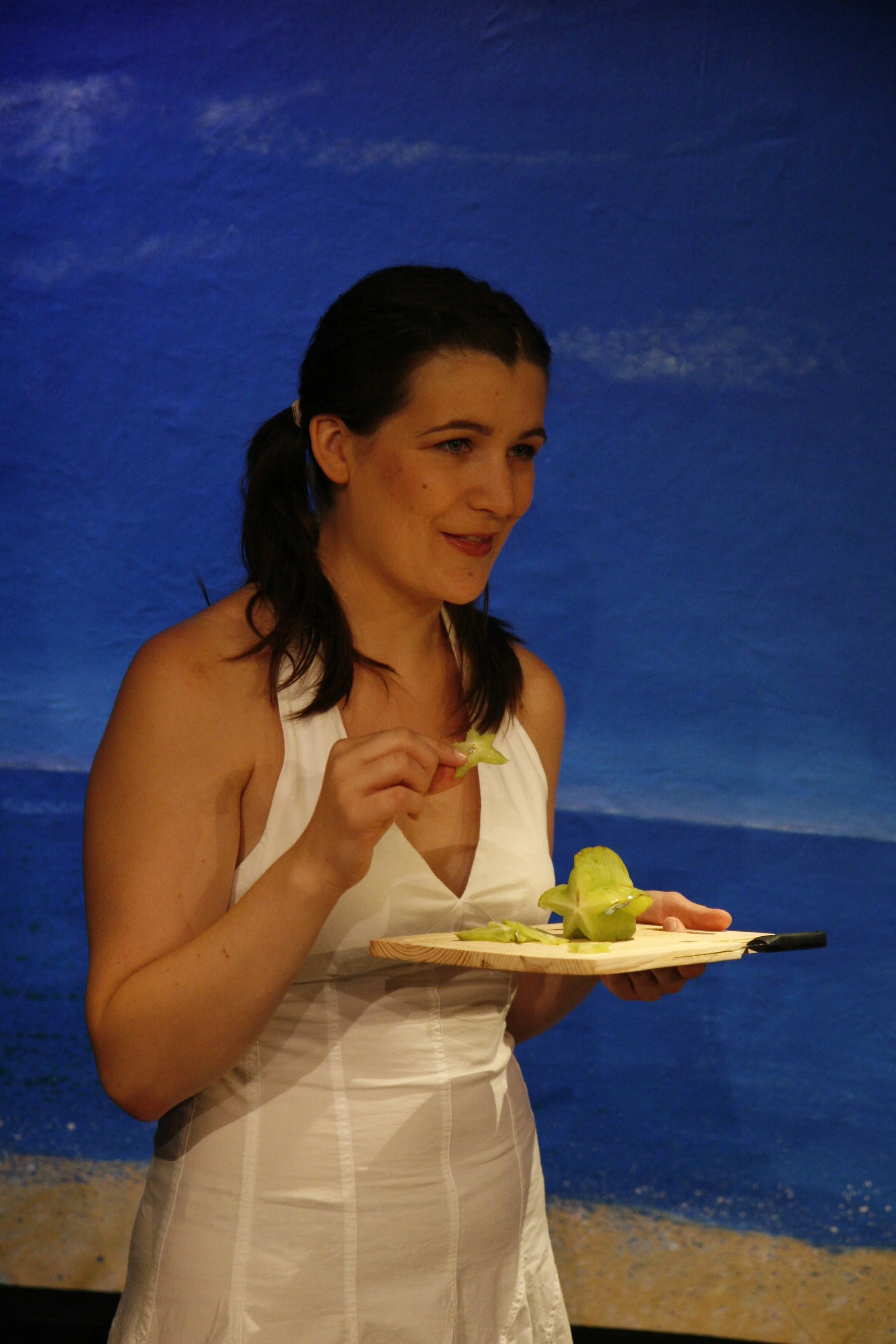A woman wearing a white sleeveless dress, is holding a wooden serving board with starfruit slices and a starfruit in her hand, standing against a backdrop of a blue ocean scene.