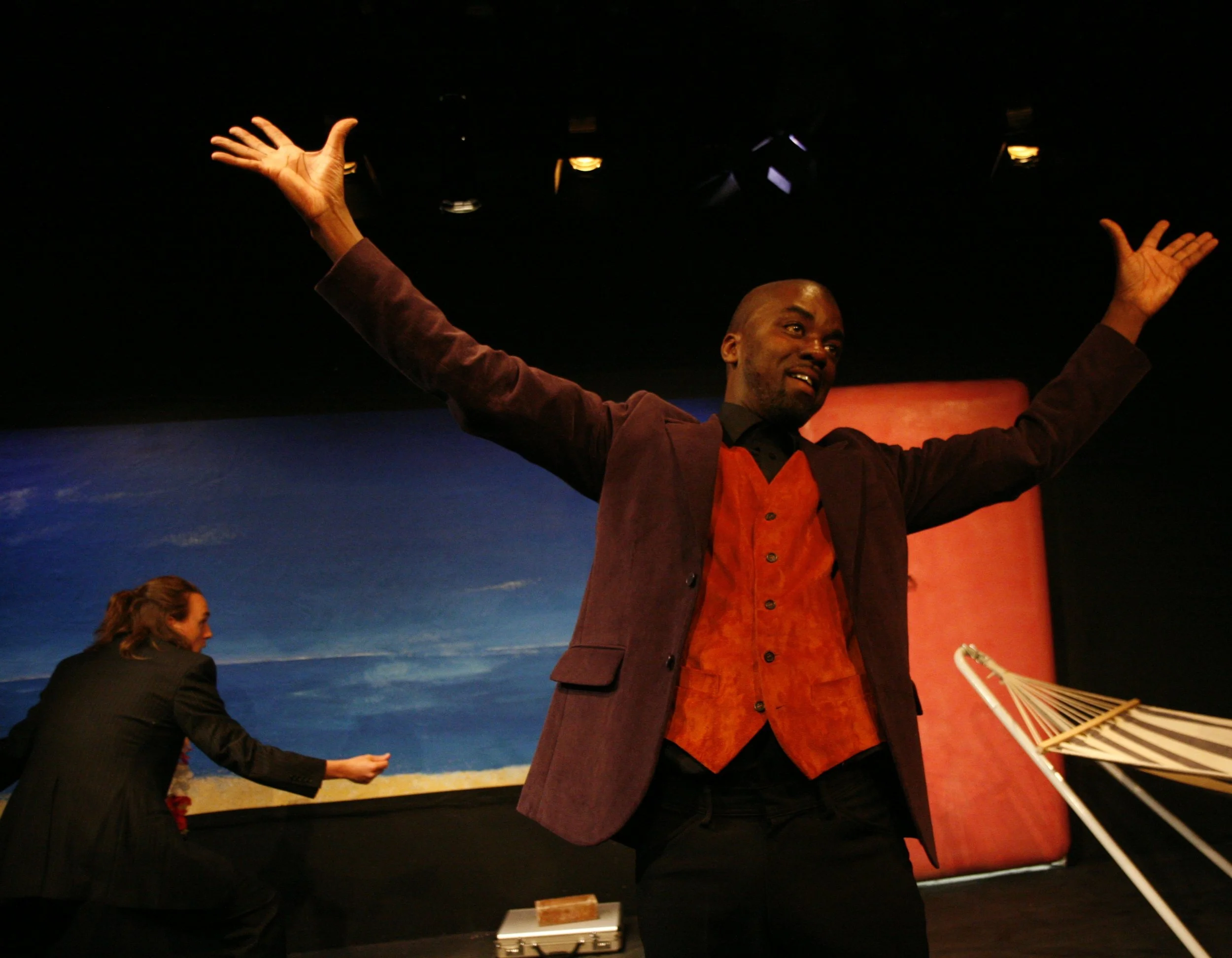A man with arms raised and smiling on stage, with a man in the background reaching out. Stage decor includes a sky and beach scene.