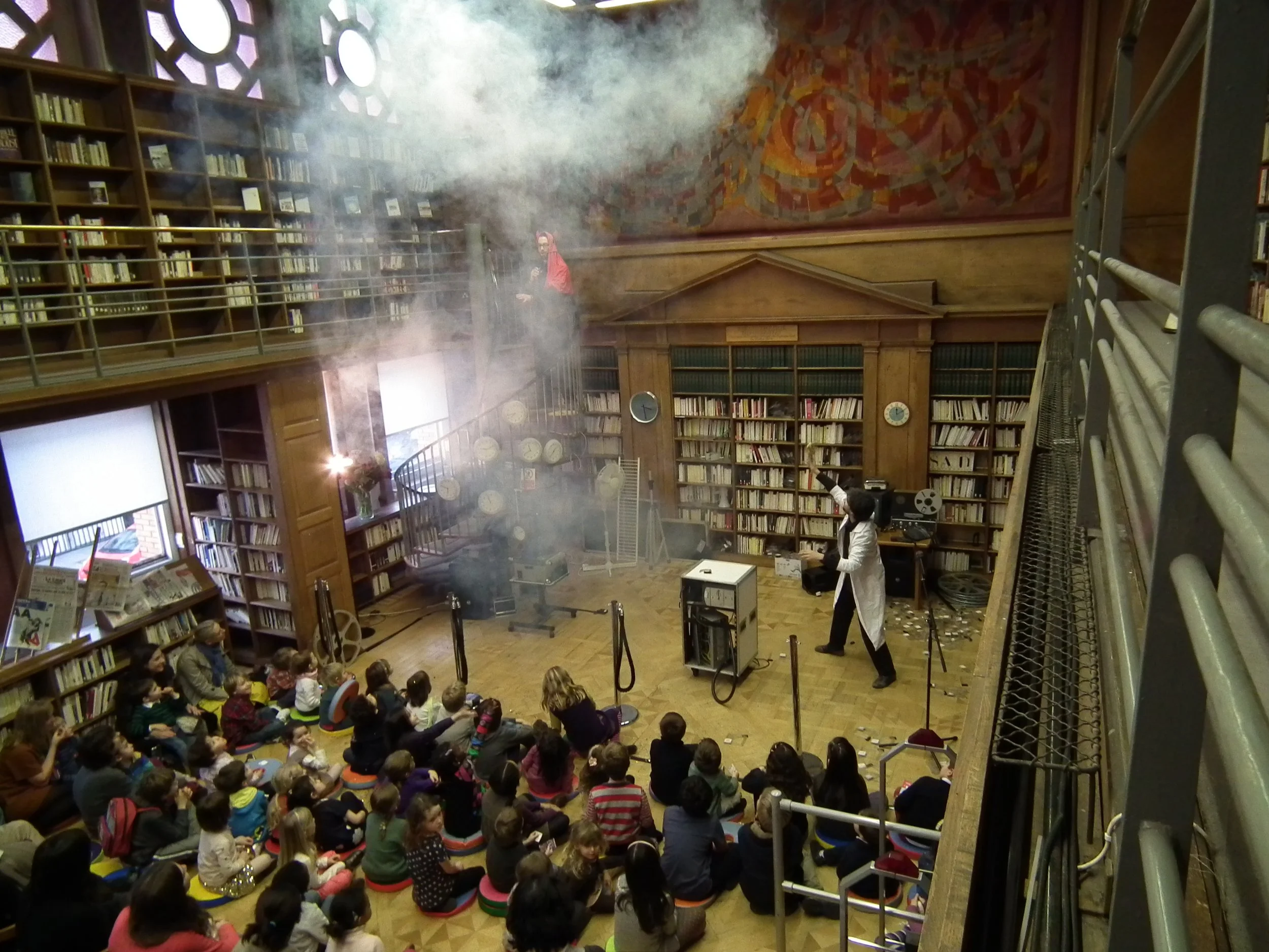 Children seated on colorful cushions in a library watching performance involving smoke and sparks, with a scientist in a white lab coat and a person on a balcony behind a cloud of smoke.