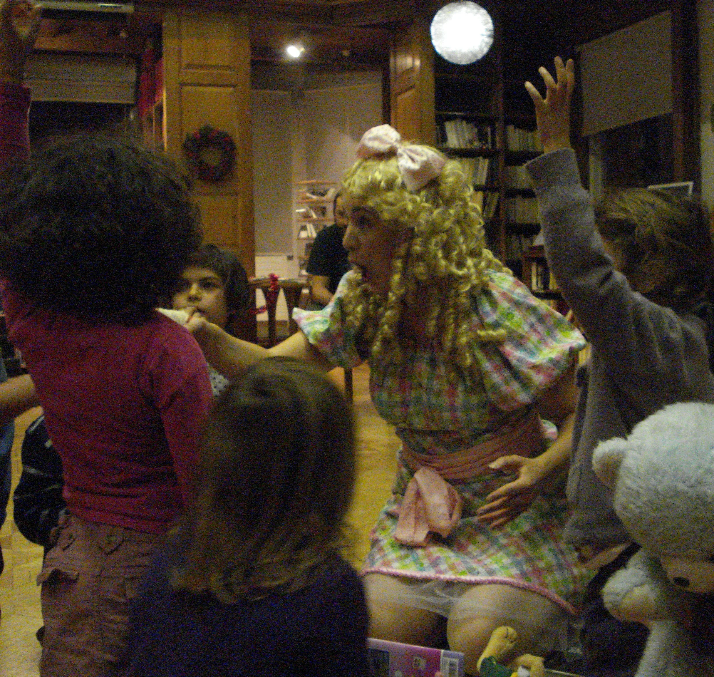 Une femme aux cheveux blonds bouclés, portant un nœud rose et une robe à carreaux colorée, interagit avec plusieurs enfants dans une pièce décorée pour Noël, avec une couronne au mur et un éclairage chaleureux.