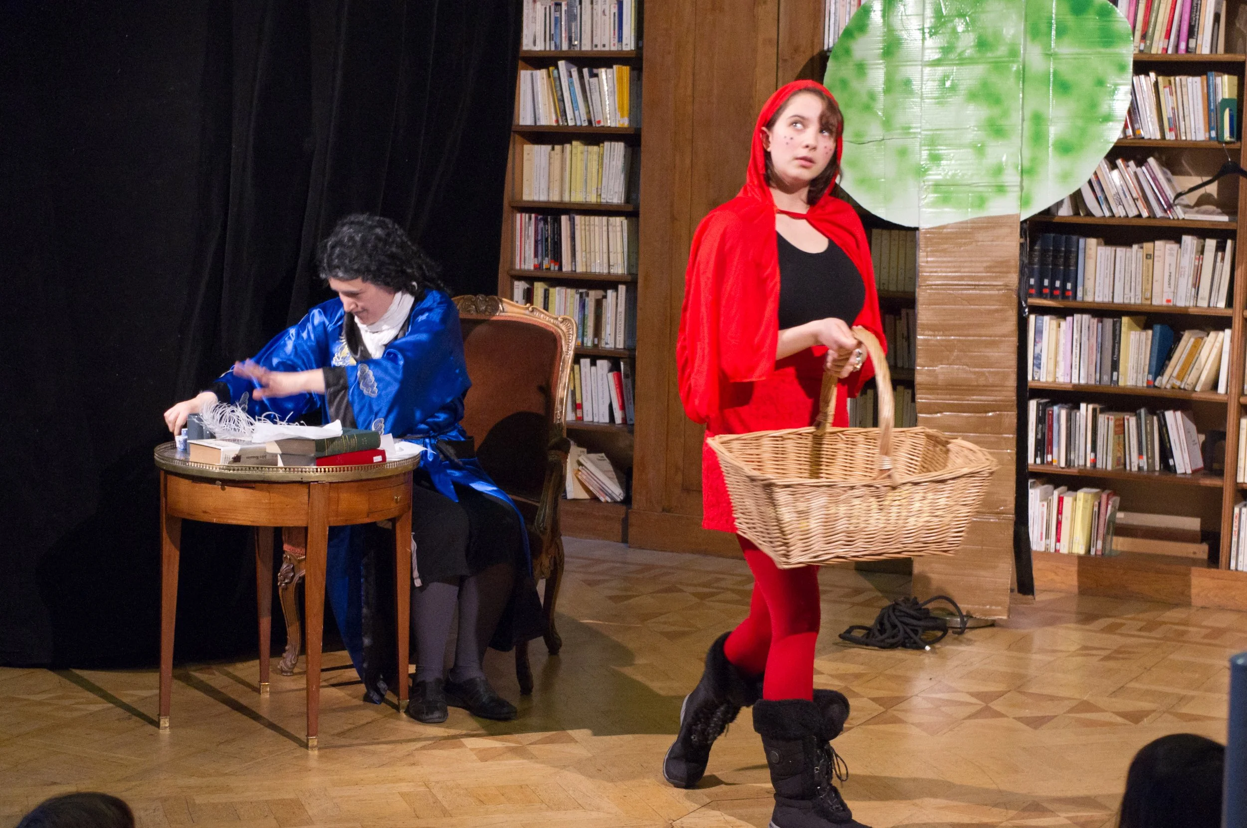 A theatrical performance with two women onstage. One is sitting at a small wooden table, dressed in a blue costume, writing something. The other, dressed as Little Red Riding Hood, stands nearby.