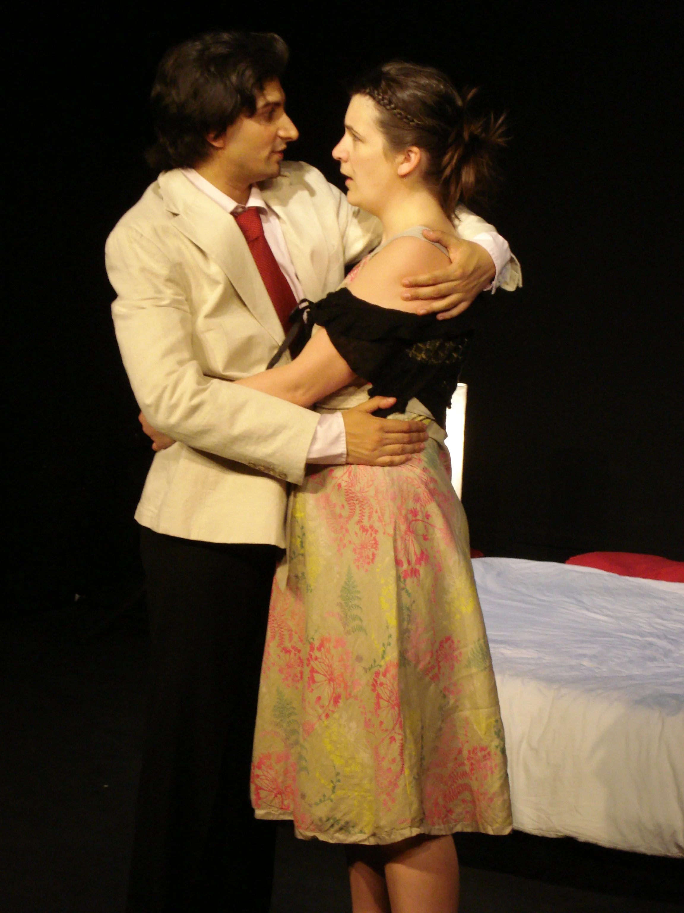 A man and woman are standing close, embracing each other and looking into each other's eyes. The man is wearing a light-colored suit and red tie, and the woman is in a black off-shoulder top and a floral skirt. There is a bed with white and red bedding in the background.