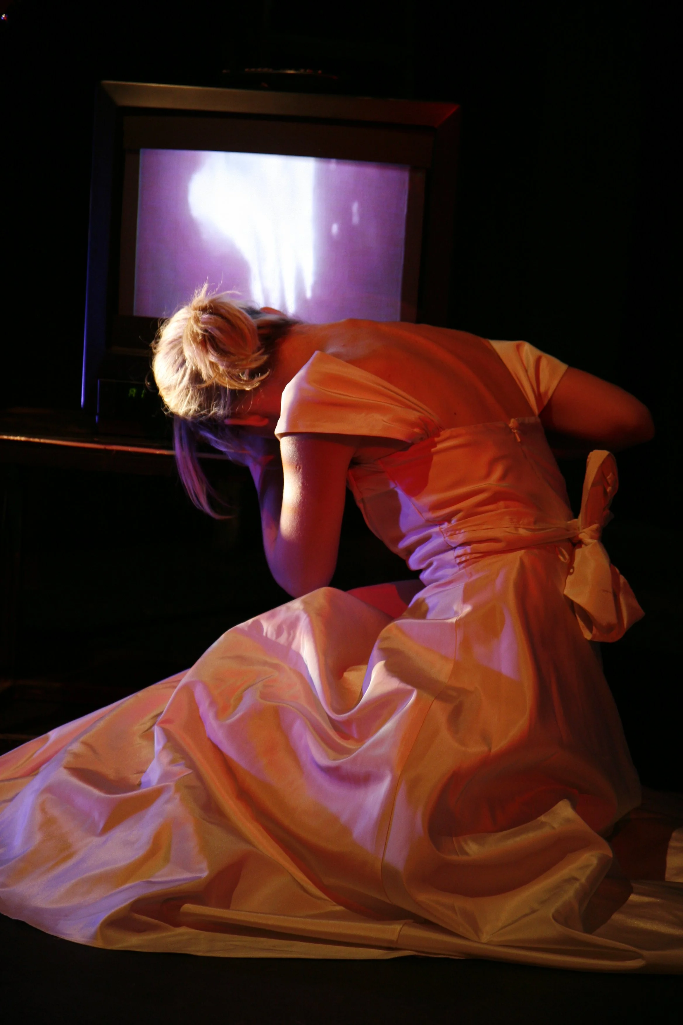 A woman in a dress kneeling on the floor with her head resting on her arm in front of a small television