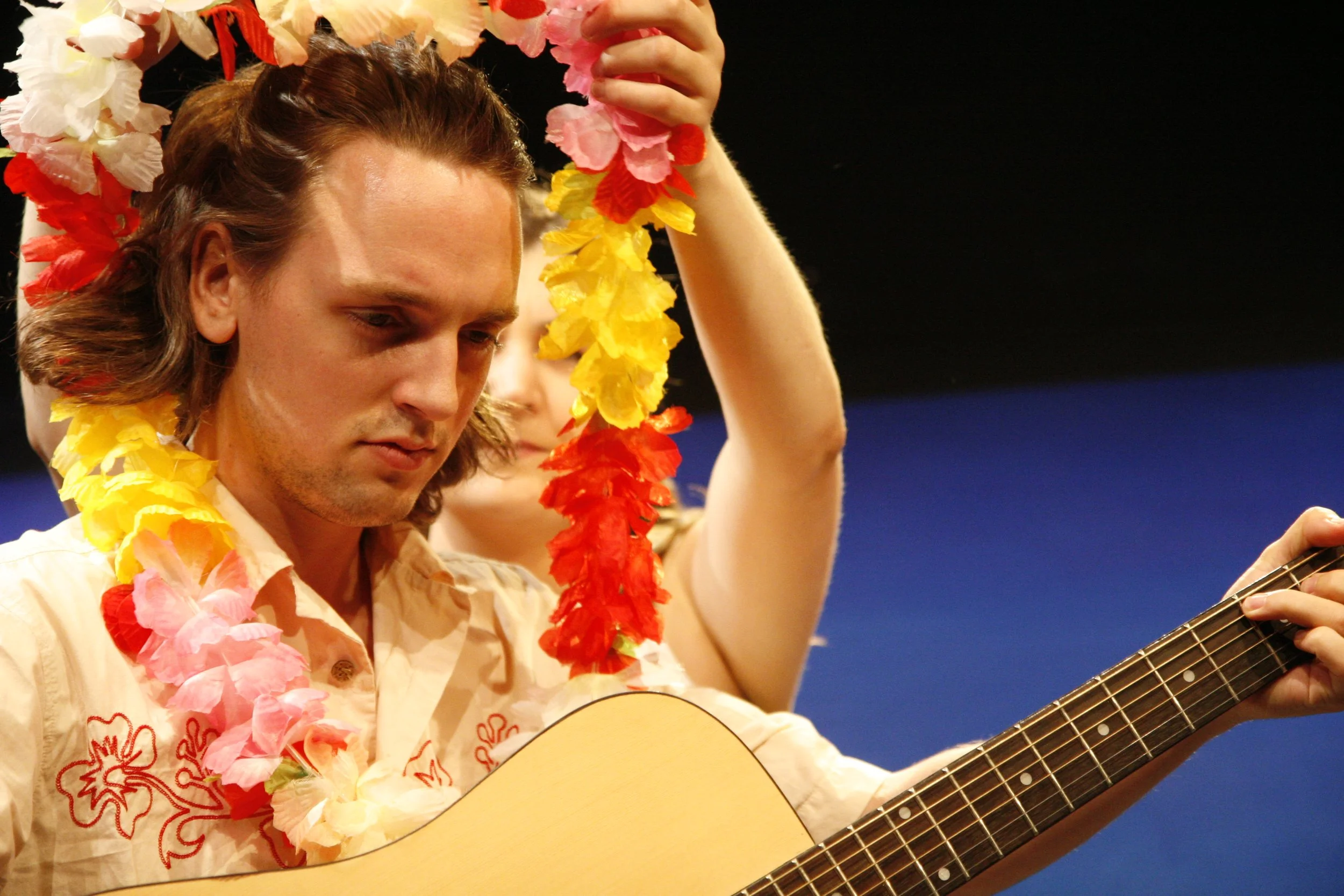 Man playing acoustic guitar wearing a floral Hawaiian shirt and a colorful Hawaiian lei, with a focused expression