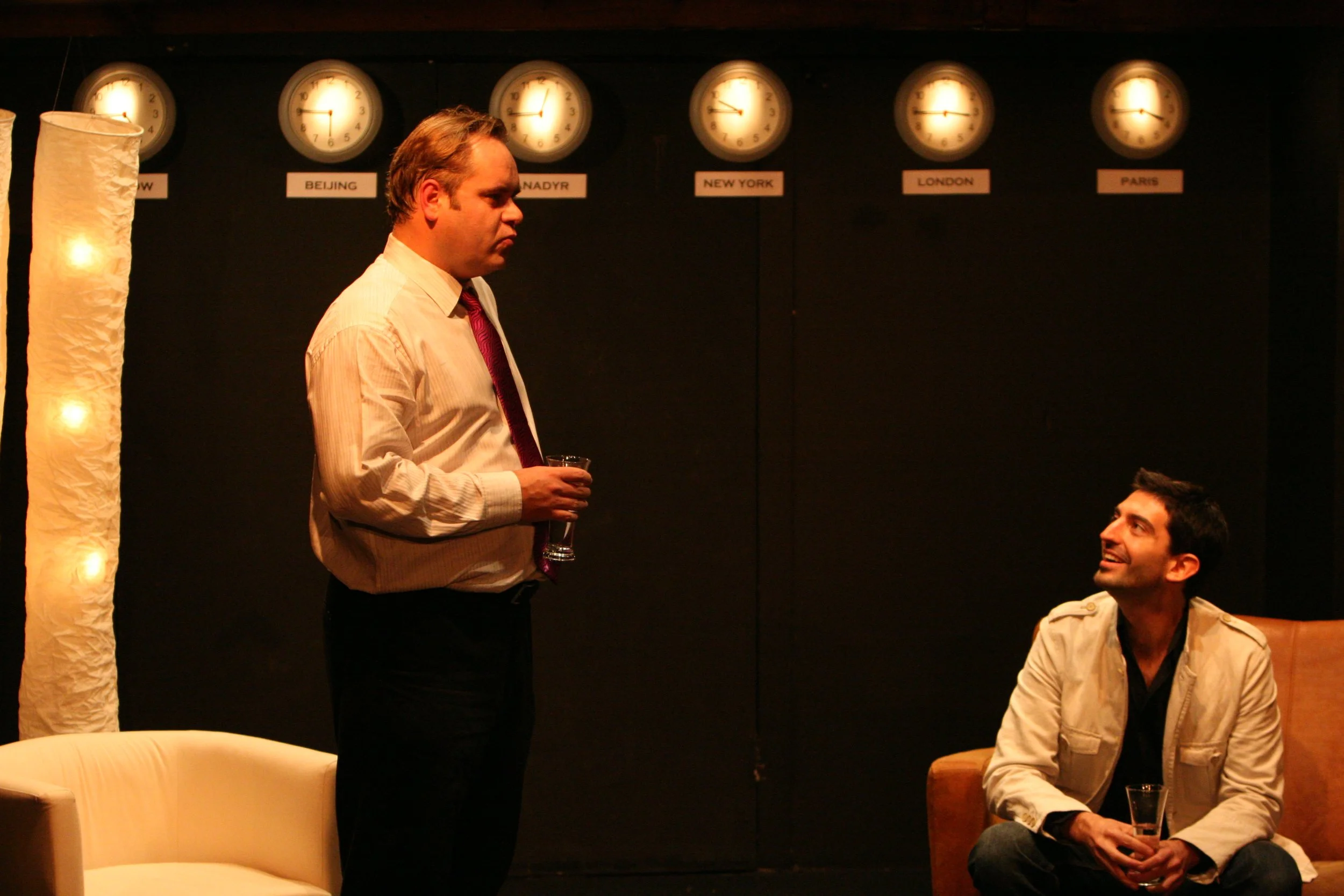 Two men having a conversation in a room with world clocks on the wall showing different international times, labeled with cities like Beijing, New York, London, and Paris.