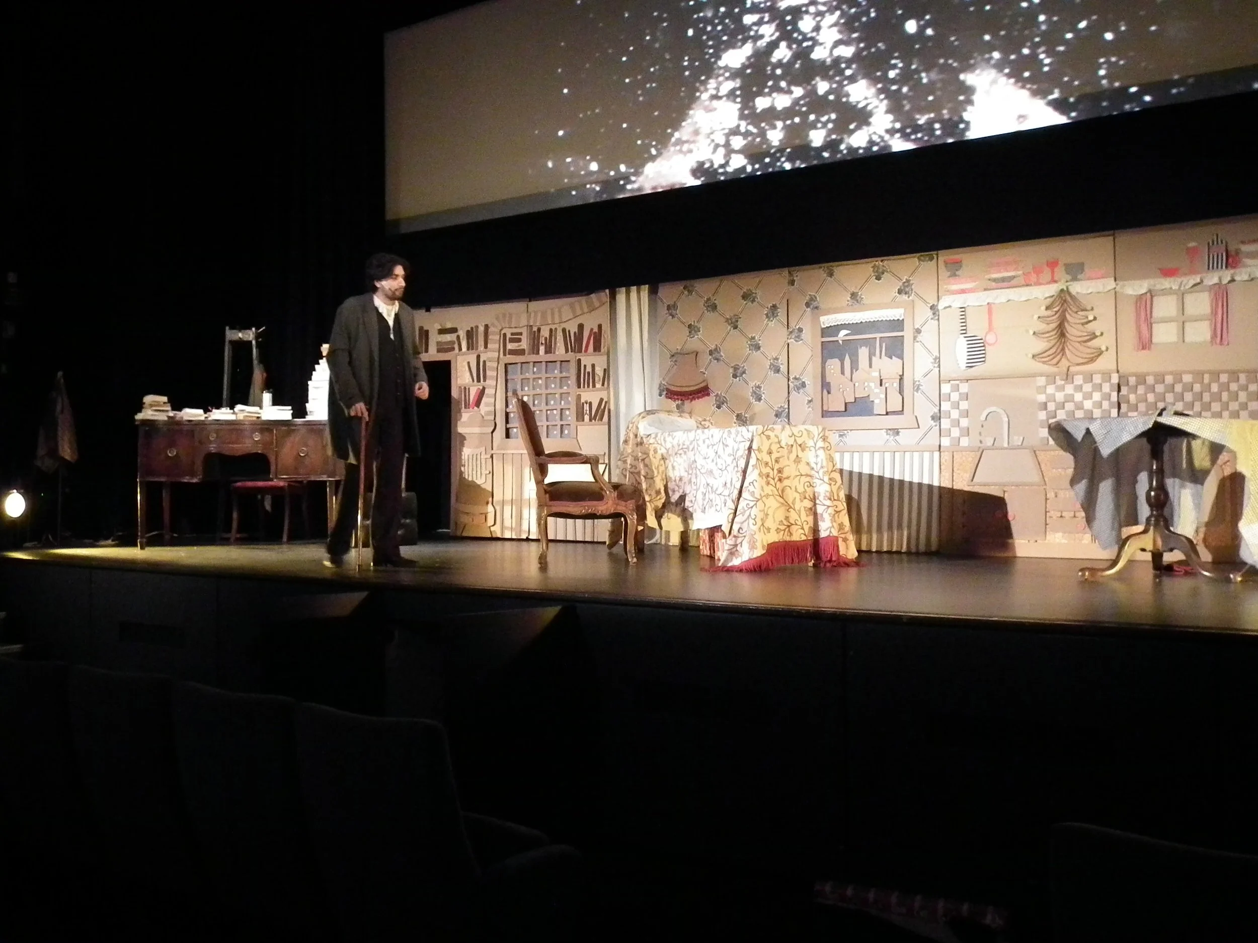 Stage set with vintage-style furniture, painted backdrop of a cozy room, a man dressed in dark clothing holding a cane, and a large screen above displaying abstract patterns.