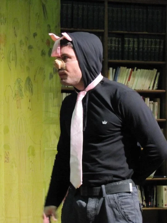 Person dressed as a pig, wearing a pink headband with pig ears, glasses, a fake snout, a black hoodie with the hood up, a white tie, and gray pants, standing in front of a yellow wall decorated with tree drawings and a bookshelf.