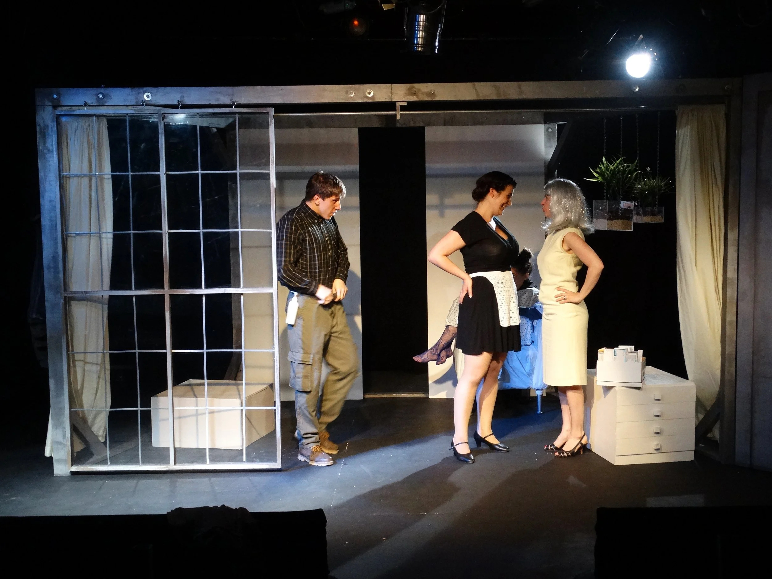 A scene from a play with four actors on stage. The set includes a large window frame, a small white chest of drawers, and a table with potted plants. Two women are standing close together, one in a black dress with a white apron and the other in a cream-colored dress with gray hair. A man and a woman are in the background, with the man holding something and the woman partially obscured.