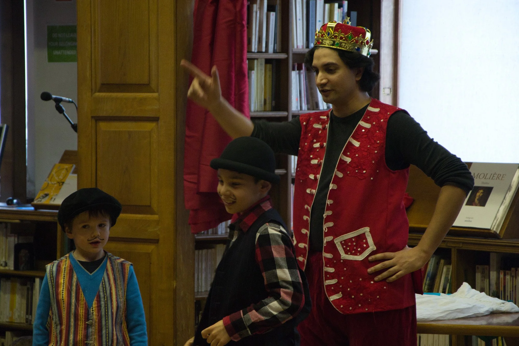 A man dressed as a king with a red vest and a crown on his head is entertaining children. A child is wearing a black beret and a painted mustache and a young boy is wearing a bowler hat.
