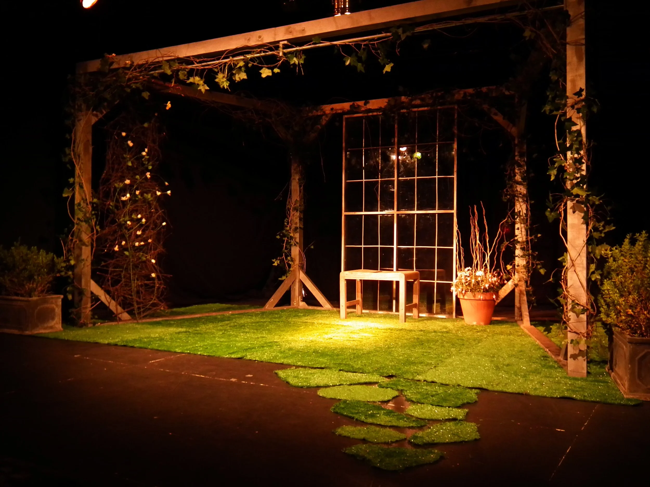 A small stage with a frame decorated with vines and plants, a grassy platform, and a large bush or flower pot on the right, illuminated by a warm yellowish light.