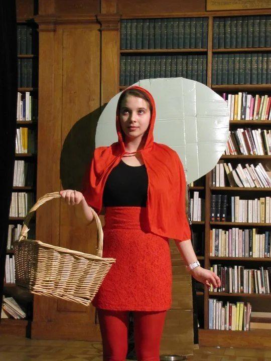 A woman dressed as Little Red Riding Hood, wearing a red cape, red skirt, black top, and red tights, standing onstage with bookshelves in the background, holding a wicker basket.