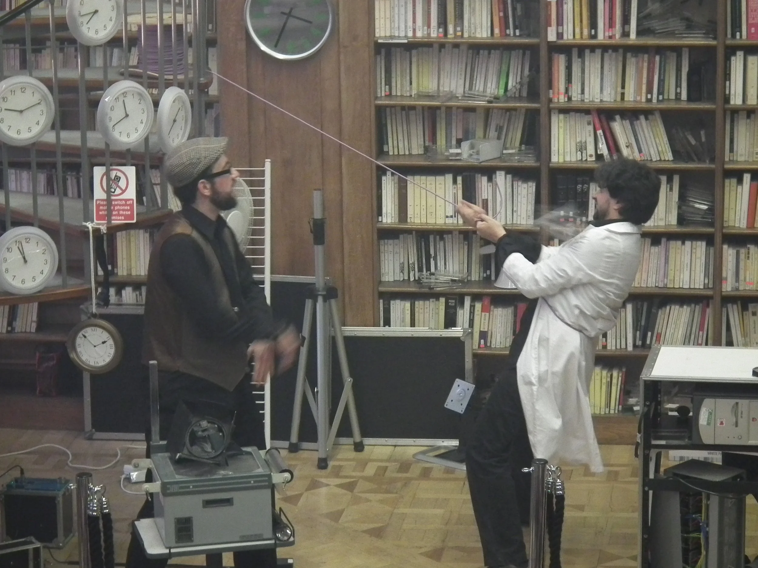 Two men onstage in a library with bookshelves, one wearing a lab coat and glasses, the other wearing a cap and vest, engaging in an experiment involving a fishing pole with a string attached, with various scientific equipment around them.