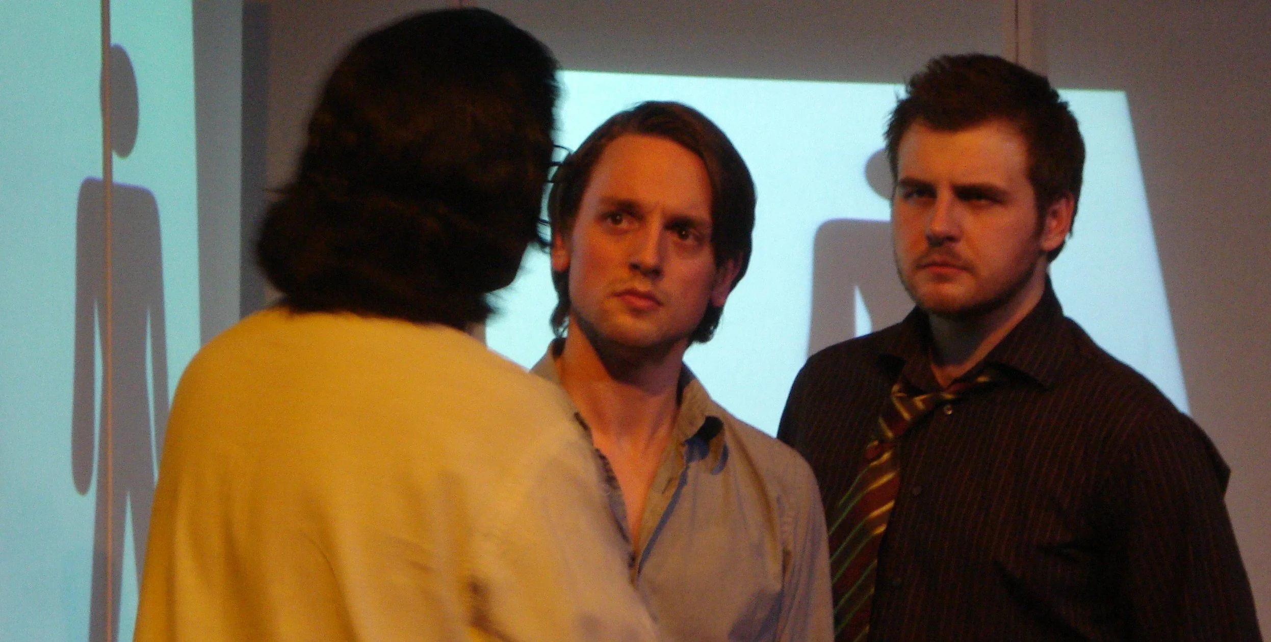 Three people in a room, two men facing someone with his back to the audience. They appear to be in a serious conversation, looking concerned and skeptical. There are silhouettes on a screen behind them.
