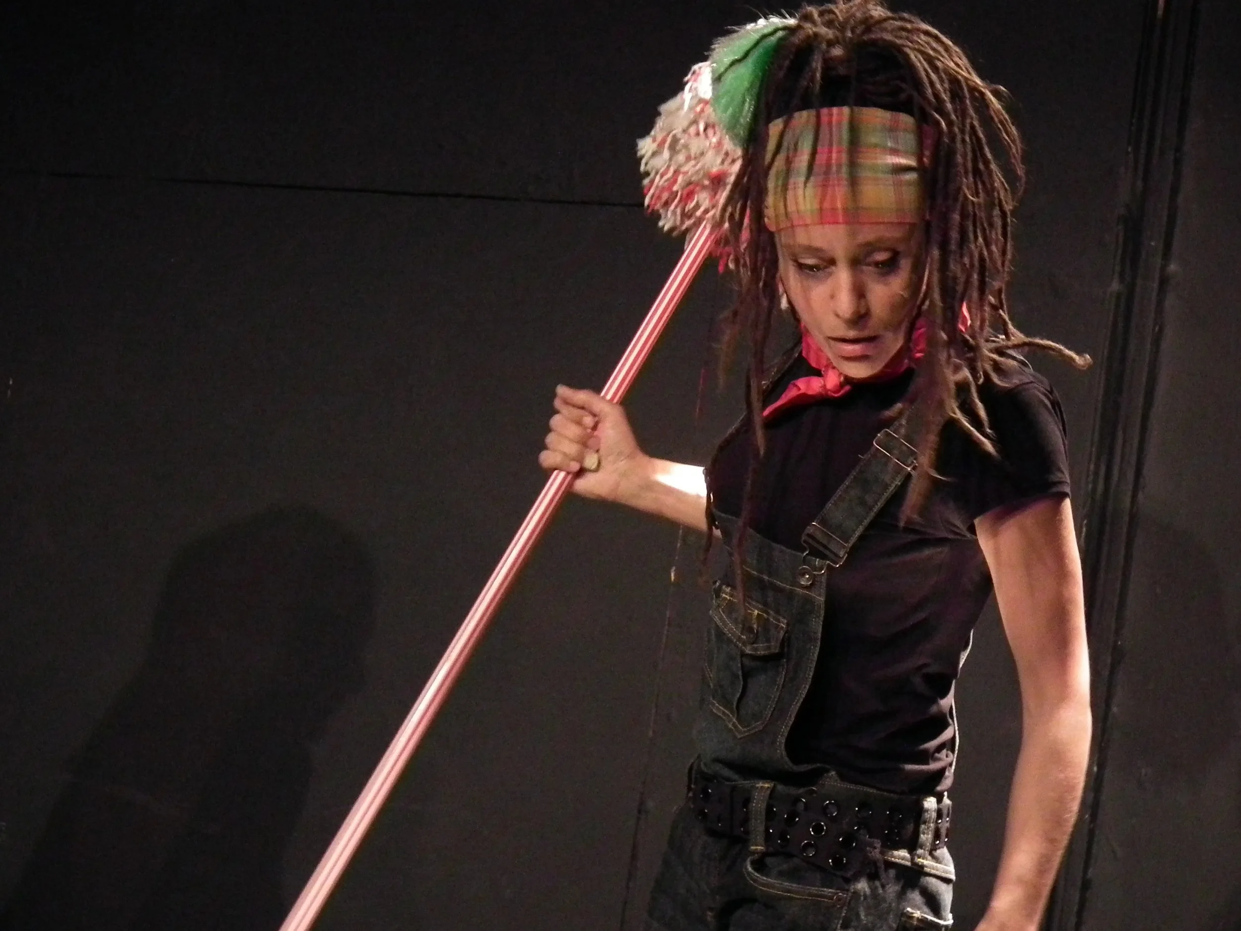A young woman wearing a plaid headband and black overalls, holds a large stick and looks down with an intense expression against a black background