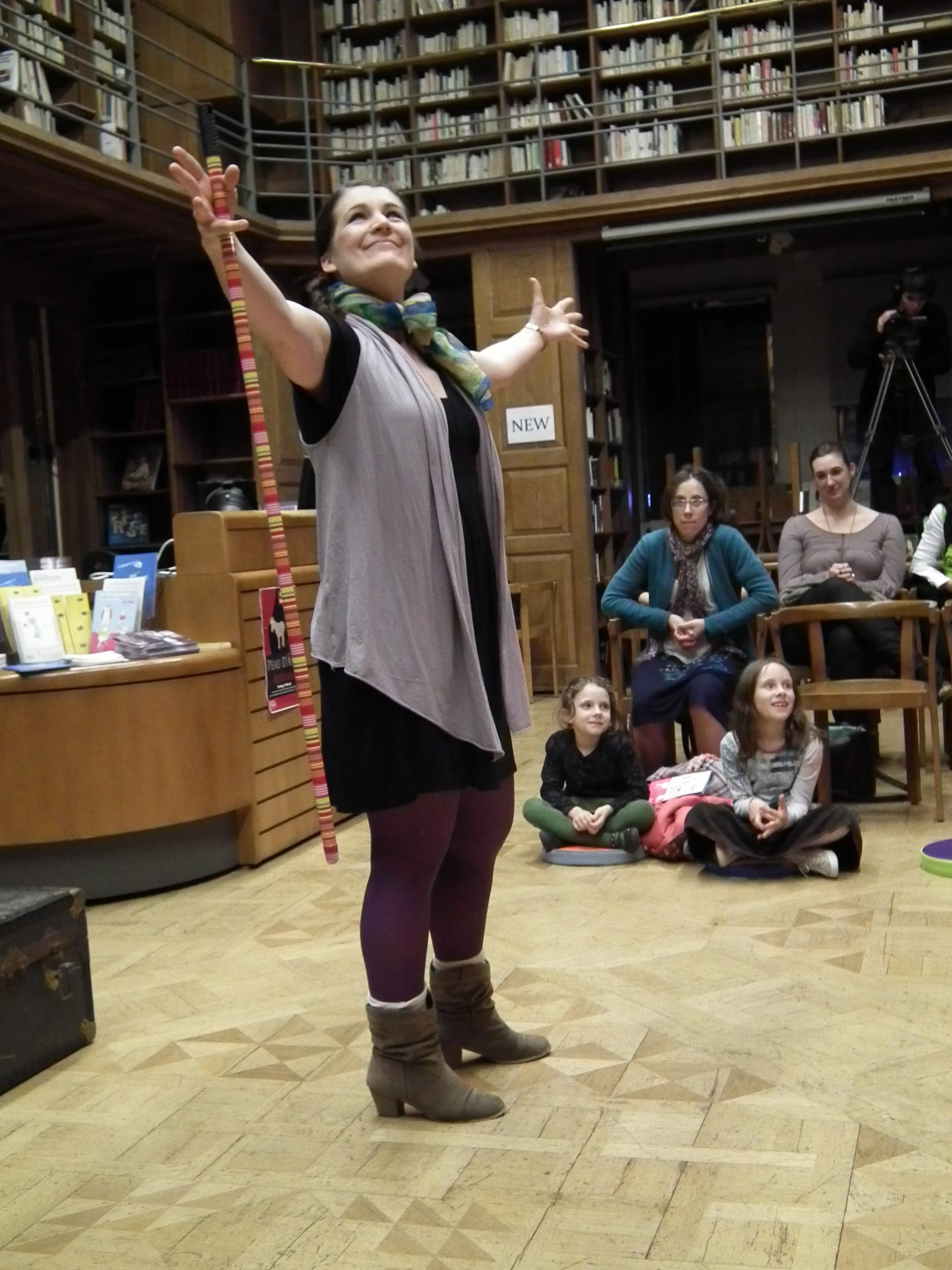A woman standing in front of an audience, gesturing with her hands. Several children and adults are seated on the floor and in chairs, watching her. There are bookshelves and a camera in the background.