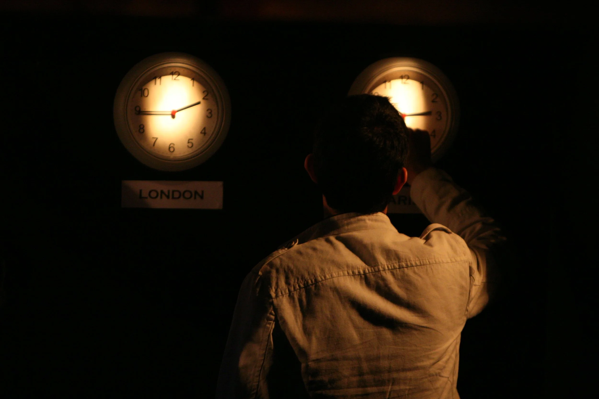 A man in a beige jacket looks at two clocks on a wall, one labeled 'London' onstage.
