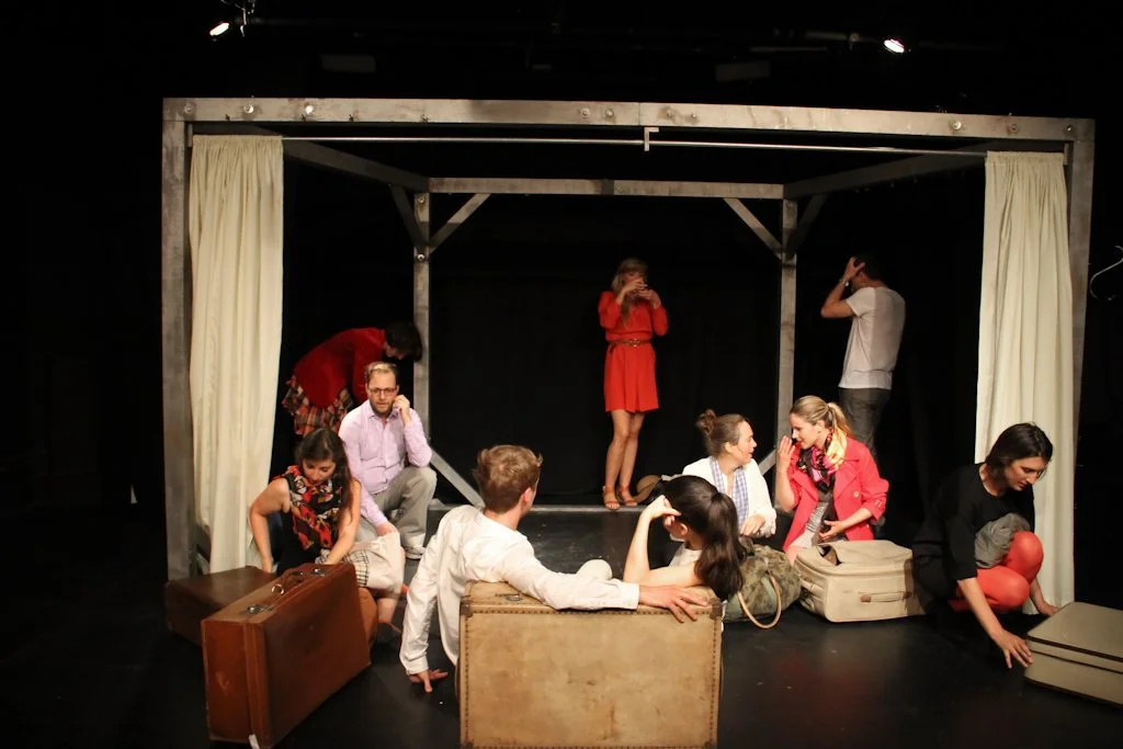 A group of people on a stage set, with some sitting and some standing, engaging in conversation; a woman in a red dress stands in the center, and others are dressed casually with luggage.